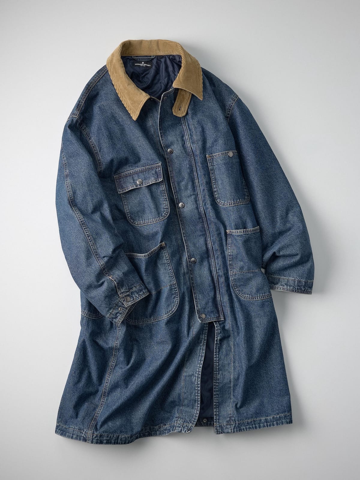 A long blue denim work coat with a tan corduroy collar, chest and hip patch pockets and a press-stud front, laid flat on a grey background