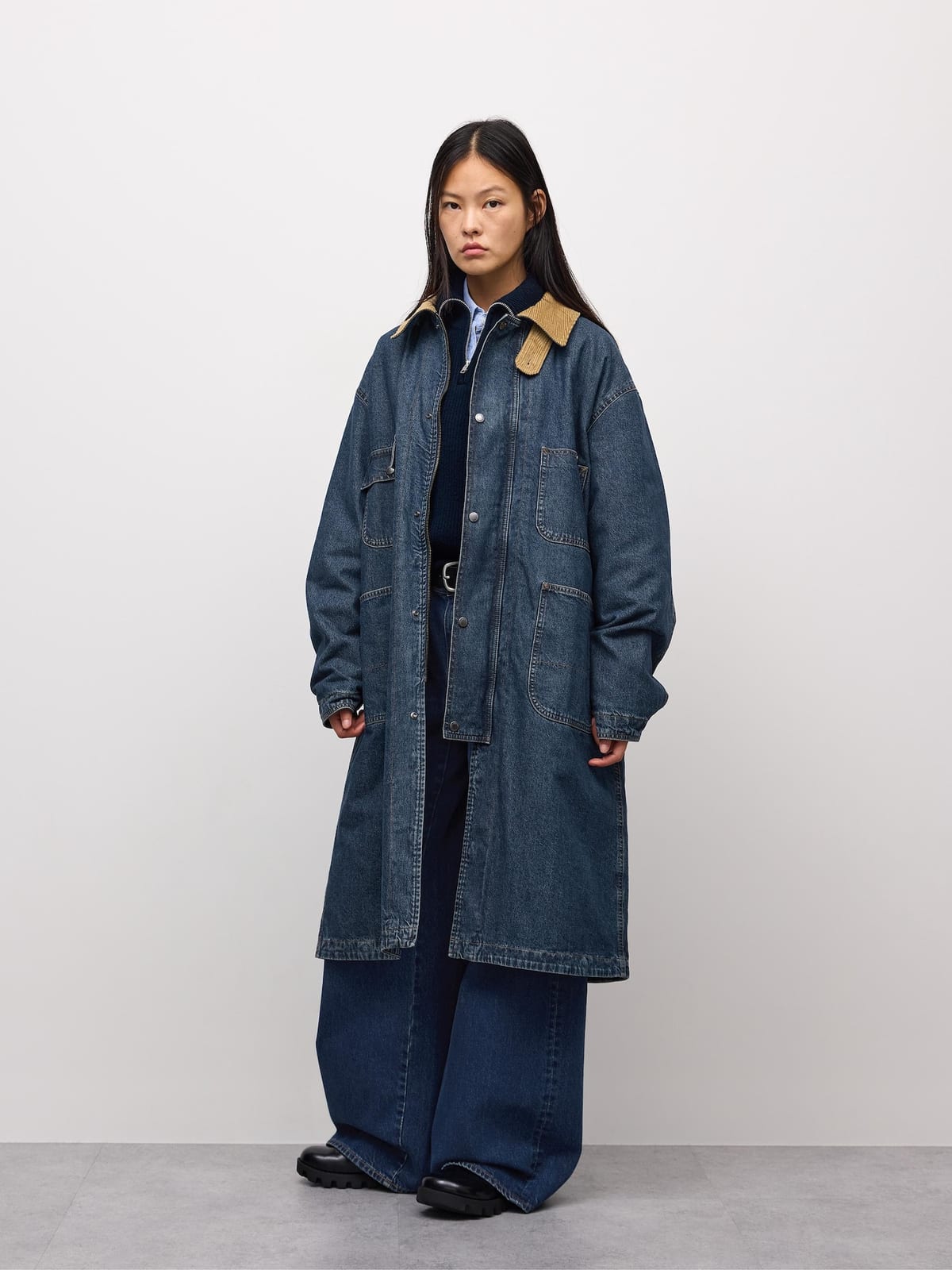 A woman with long dark hair stands against a white wall in a long blue denim work coat with a tan corduroy collar and patch pockets, over a navy knit and very wide dark jeans