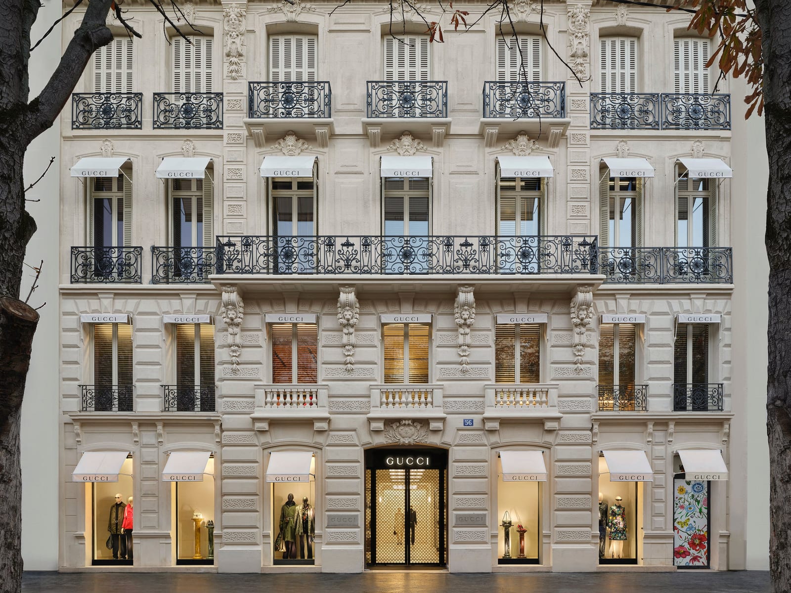 The cream stone facade of Gucci's flagship at 56 Avenue Montaigne in Paris, its upper floors lined with wrought-iron balconies and white awnings printed GUCCI, with a gold-lit entrance and mannequins in the ground-floor windows