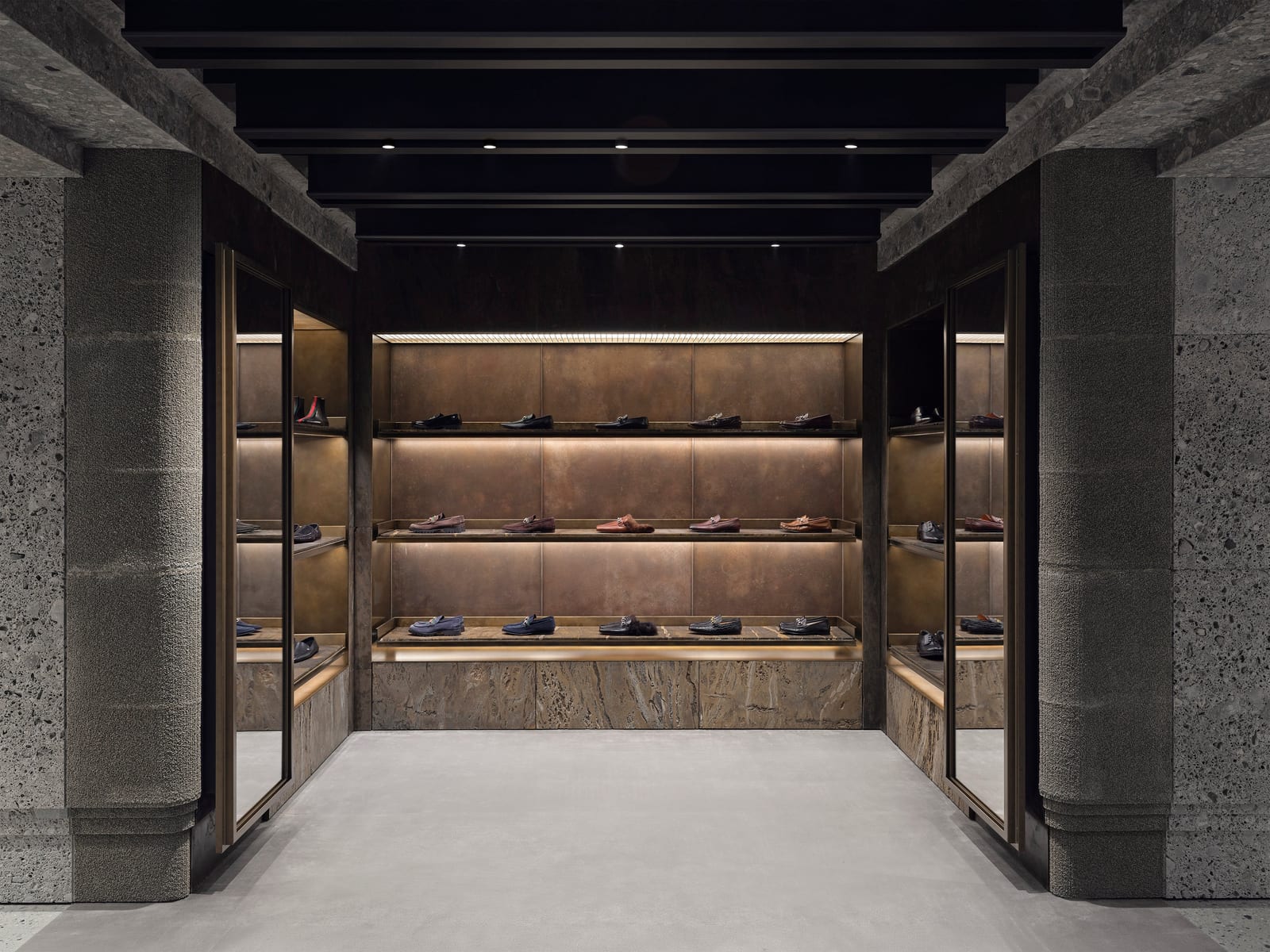 A room at Gucci's Avenue Montaigne store lined with rough grey stone pillars and a dark beamed ceiling, three lit walls of shelves holding men's loafers and dress shoes
