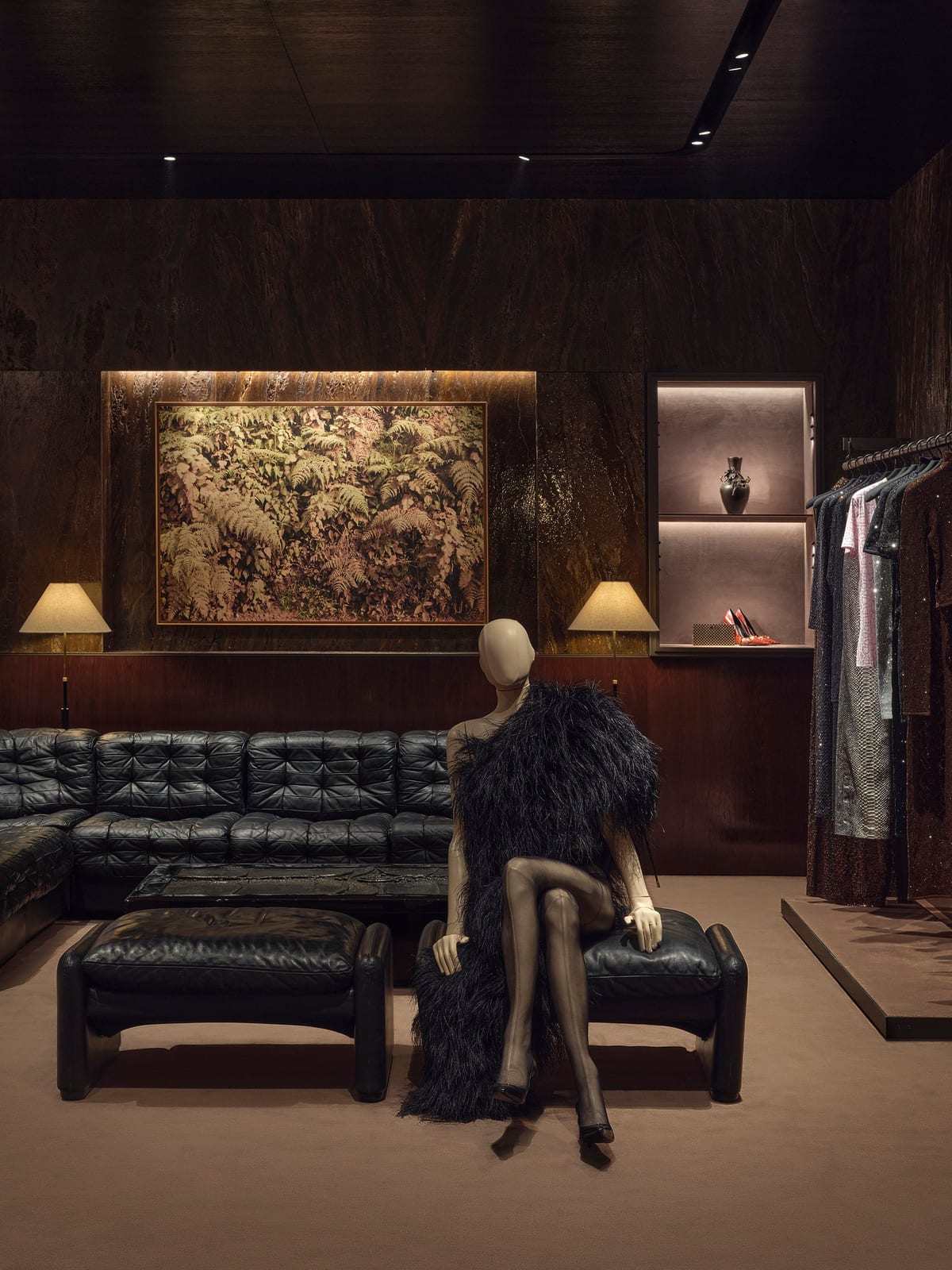 A dark-panelled salon at Gucci's Avenue Montaigne store with a long black leather sofa, a large picture of ferns between two lamps, a mannequin in a black feathered piece and a rail of sparkling dresses