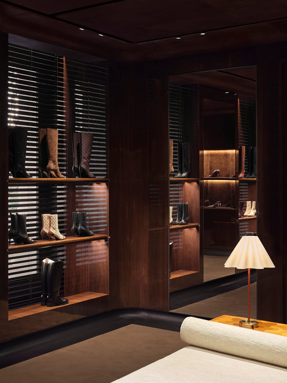 A Gucci shoe room panelled in dark walnut with slatted blinds, shelves of women's boots including brown monogram knee-highs, a mirror wall and a pleated table lamp