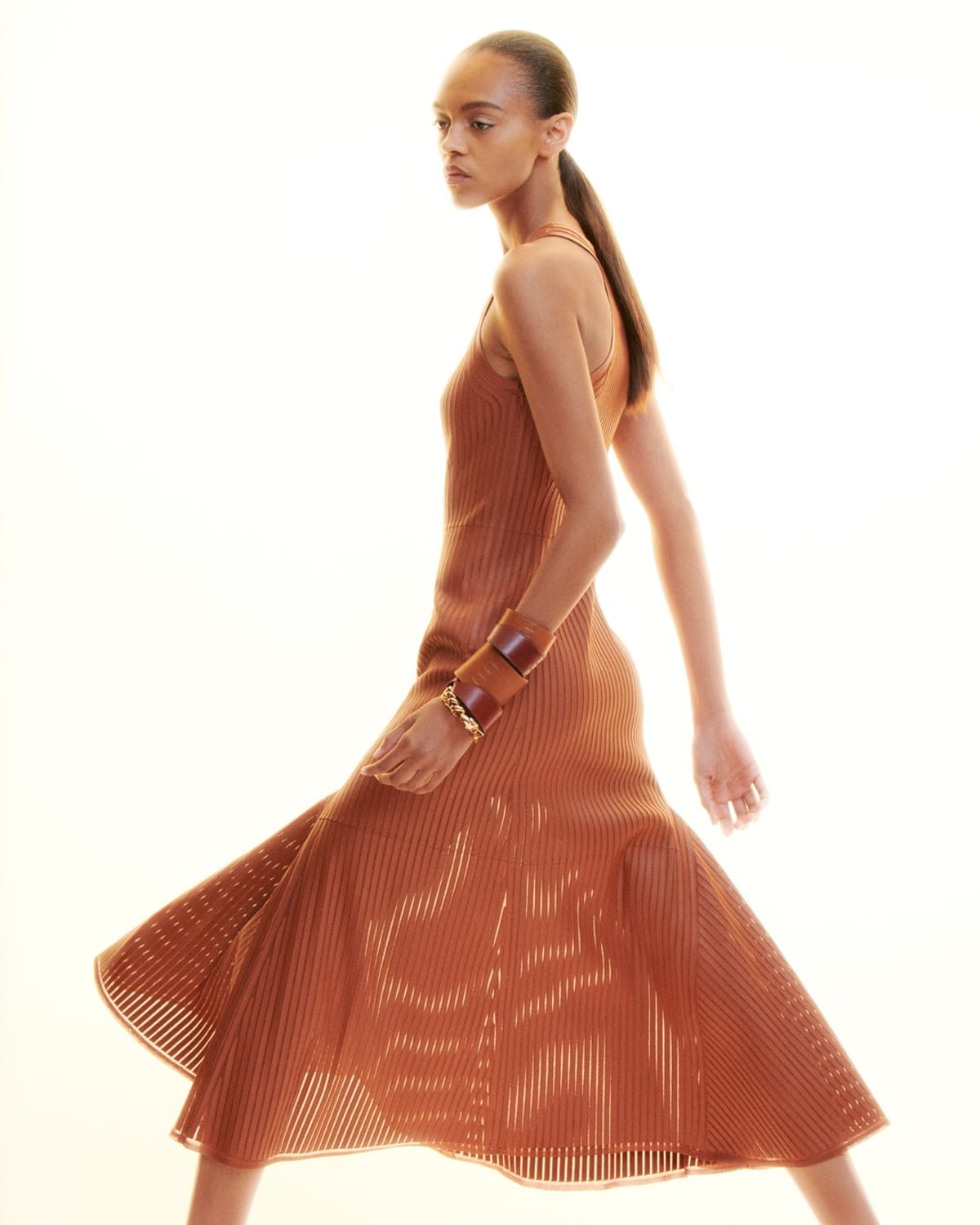 A Hermès Spring 2027 look: a sleeveless rust-coloured ribbed knit dress with a flared hem, worn with leather bracelets