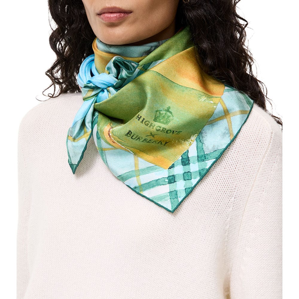 A woman in a cream jumper wearing the Birkhall silk scarf knotted at the neck, showing the green and yellow watercolour panel with the Highgrove crown and Burberry logo, and the blue check border