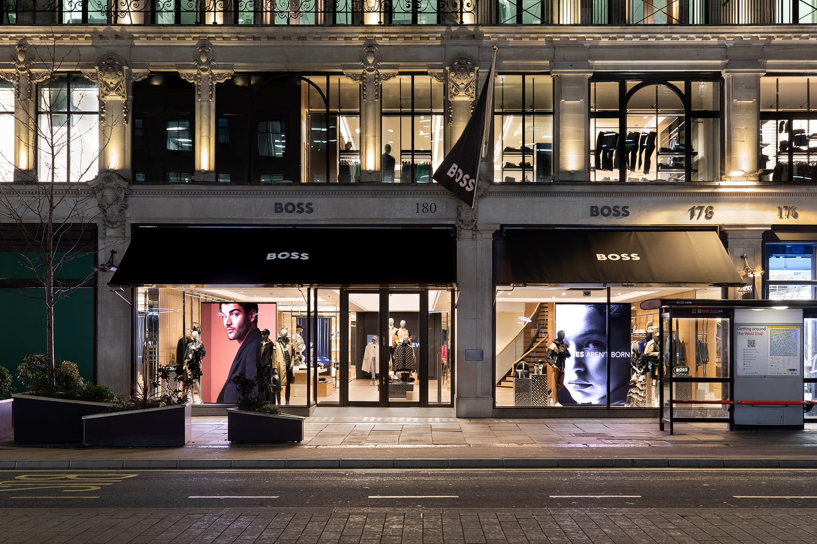 The BOSS store on London's Regent Street at night, with black BOSS awnings over lit windows, two large screens showing models' faces and a black BOSS flag on the pale stone facade, numbered 180 and 178