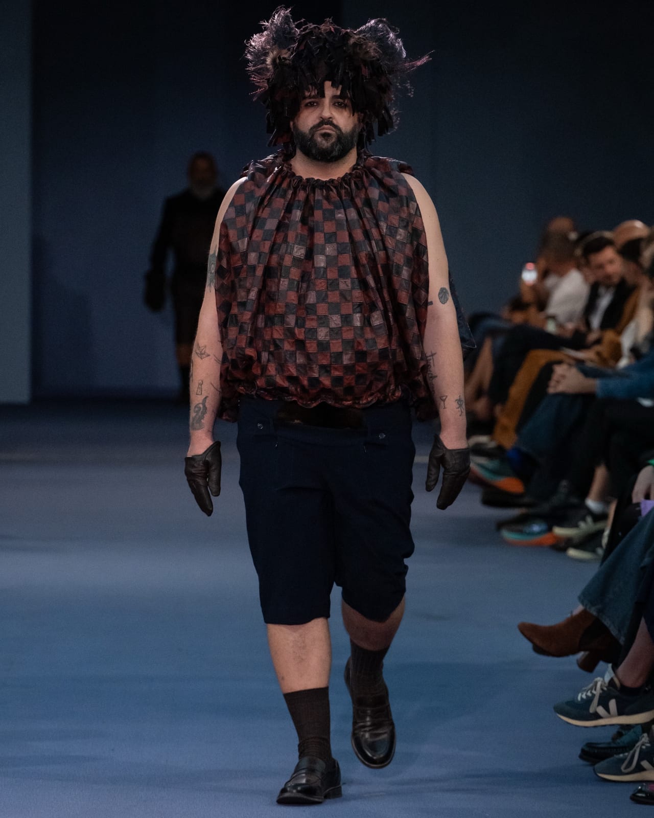 A bearded model on the runway in a gathered sleeveless top made of brown, burgundy and black leather checks, navy knee-length shorts, brown leather gloves and a shaggy headpiece of dark leather strips