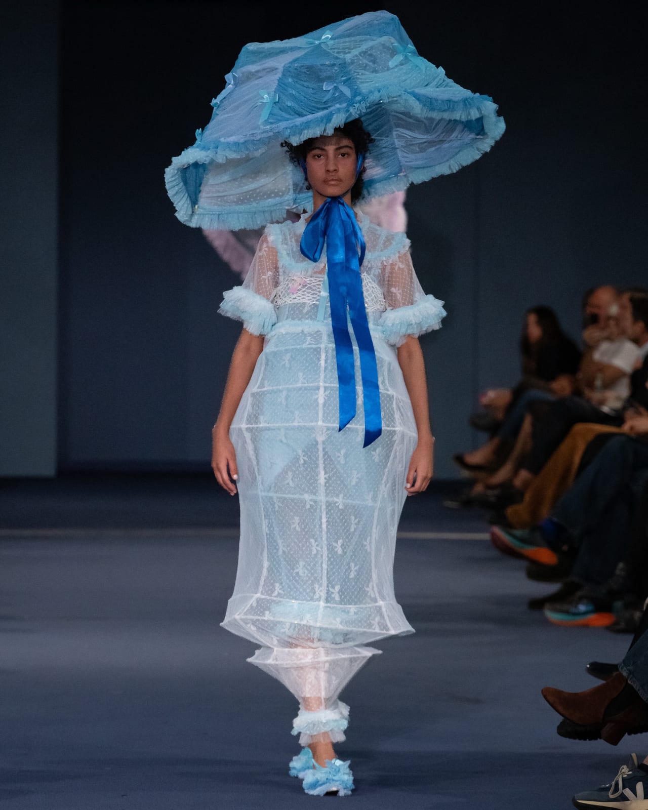 A model on a blue runway in a sheer white tulle dress built over a caged, lantern-like frame, with ruffled pale blue cuffs, a long royal blue satin ribbon at the neck and a huge ruffled pale blue hat, as the audience watches from the side
