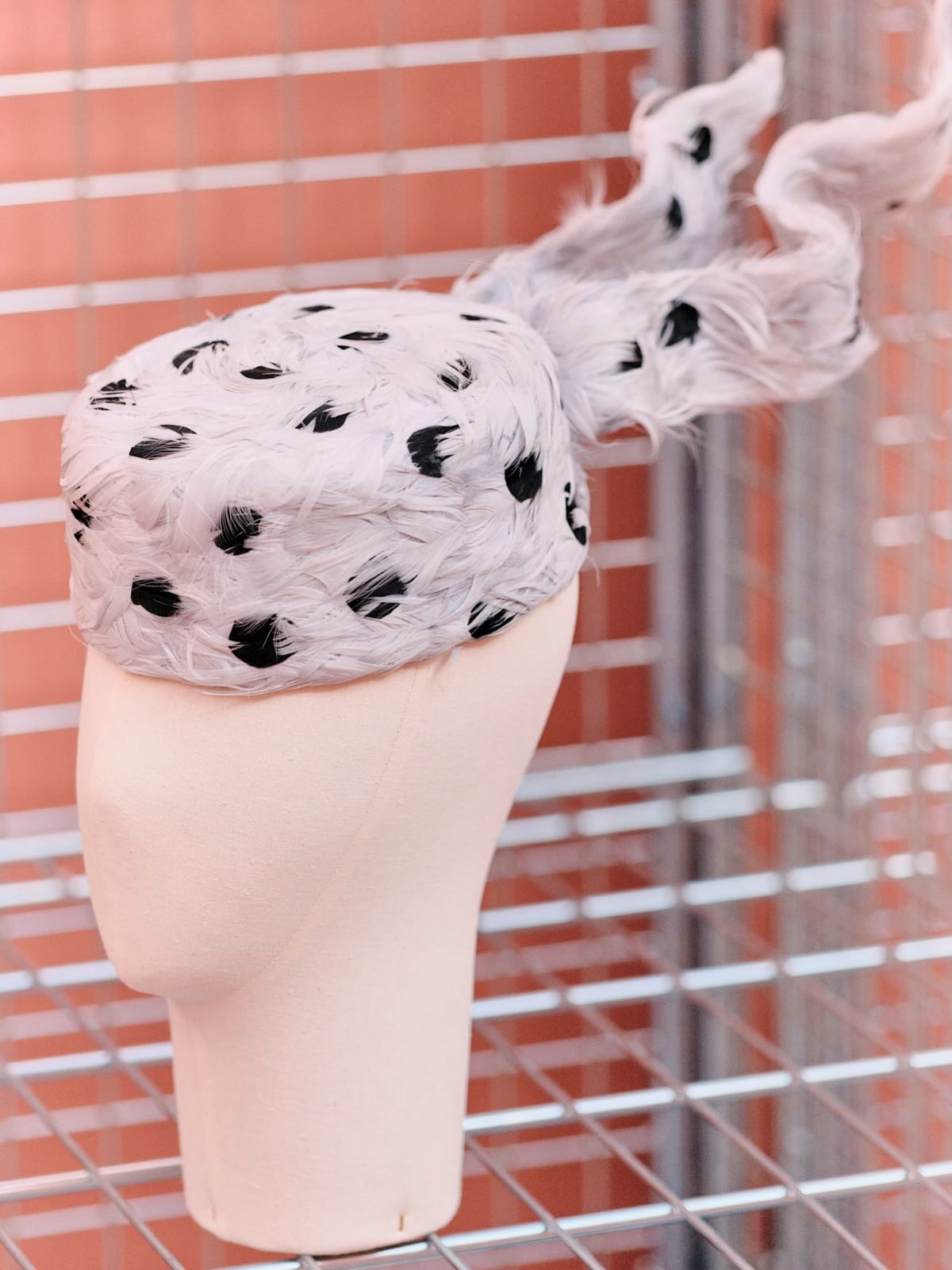 A small round hat covered in pale lilac feathers dotted with black ones, a long twisting plume of the same feathers rising from the back, on a beige mannequin head against an orange wall and steel grid