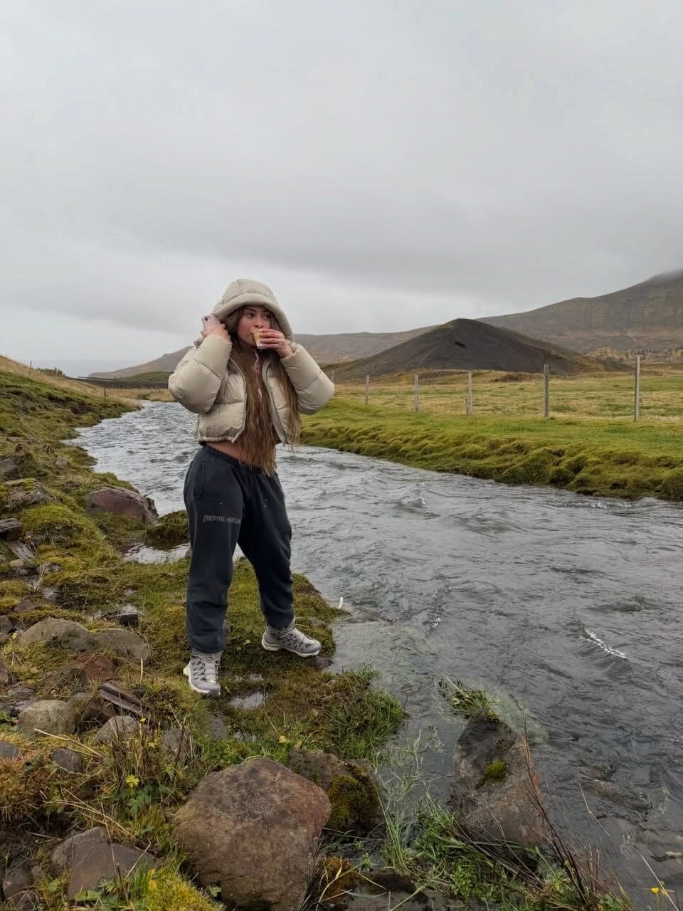 Ice Spice standing on the bank of a fast-running river in Iceland in a cream hooded puffer jacket, charcoal sweatpants and grey sneakers, with green fields and a dark hill behind her