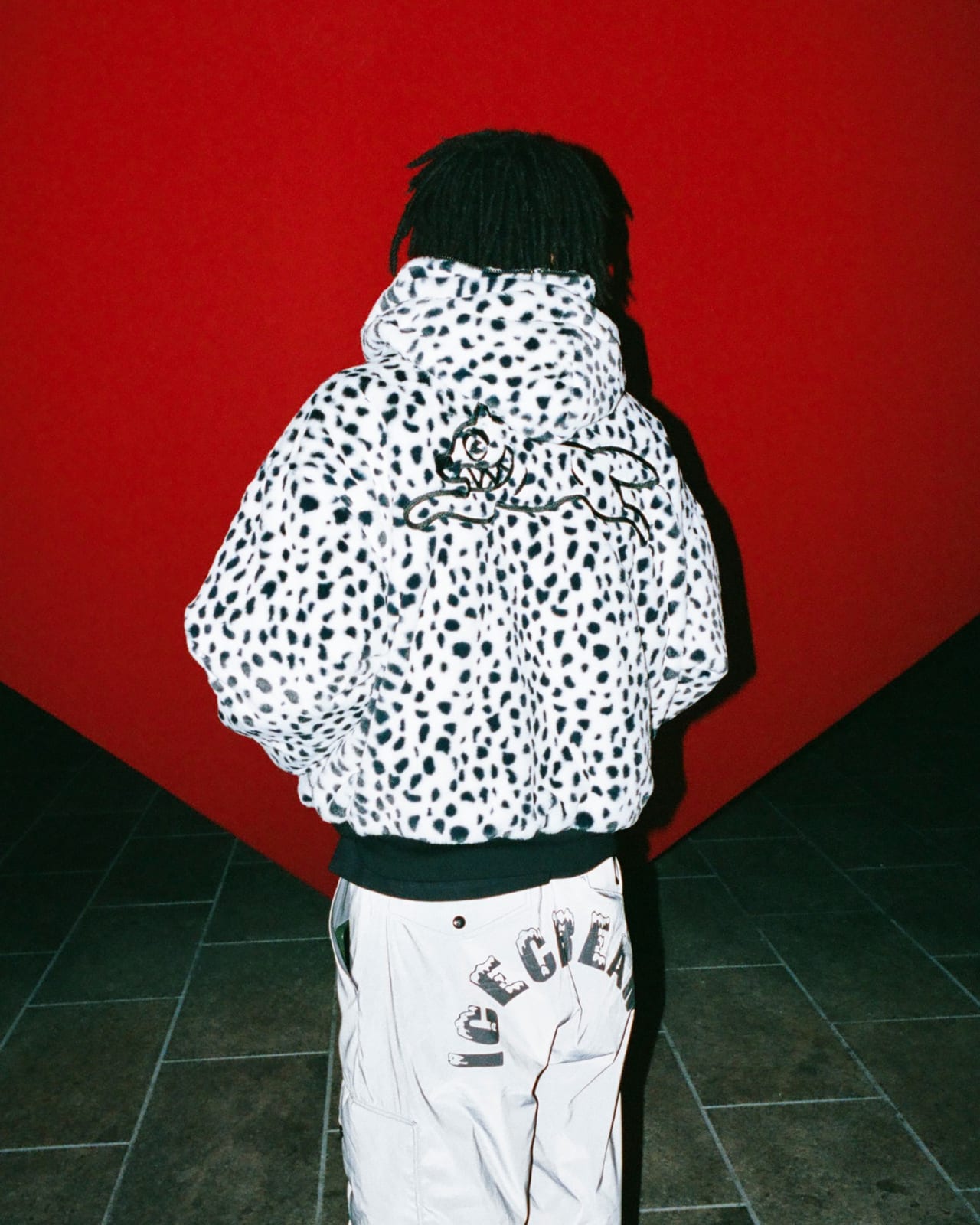 The back of a Dalmatian-print faux-fur hooded jacket with a running-dog logo embroidered between the shoulders, worn against a red wall over reflective pants lettered ICECREAM