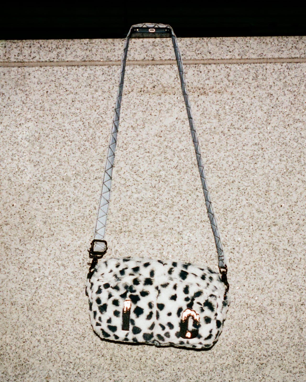 A Dalmatian-print faux-fur messenger bag with reflective I and C patches on the front and a grey strap, hung against a granite wall