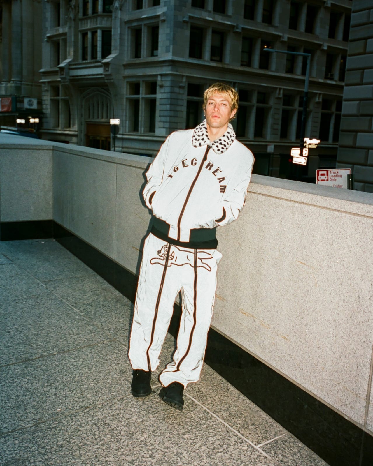 A man with cropped blond hair leaning on a granite wall on a New York street, in a white-looking reflective bomber jacket with an arched ICECREAM wordmark and a spotted faux-fur collar, over matching reflective pants with a running-dog graphic
