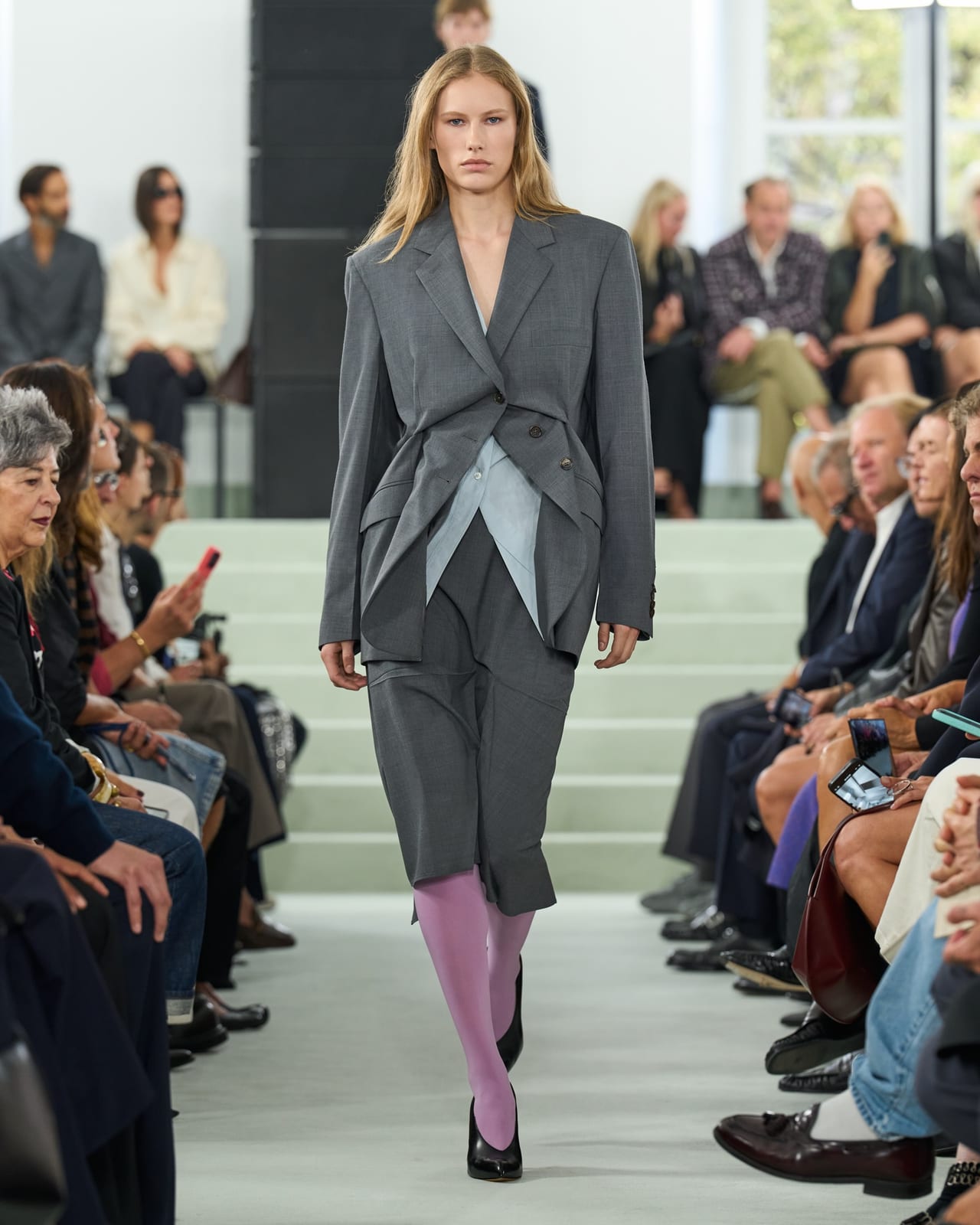 A Jil Sander Spring 2027 look: an oversized grey blazer worn open over a pale layered shirt, with a matching grey skirt, pink tights and black flats