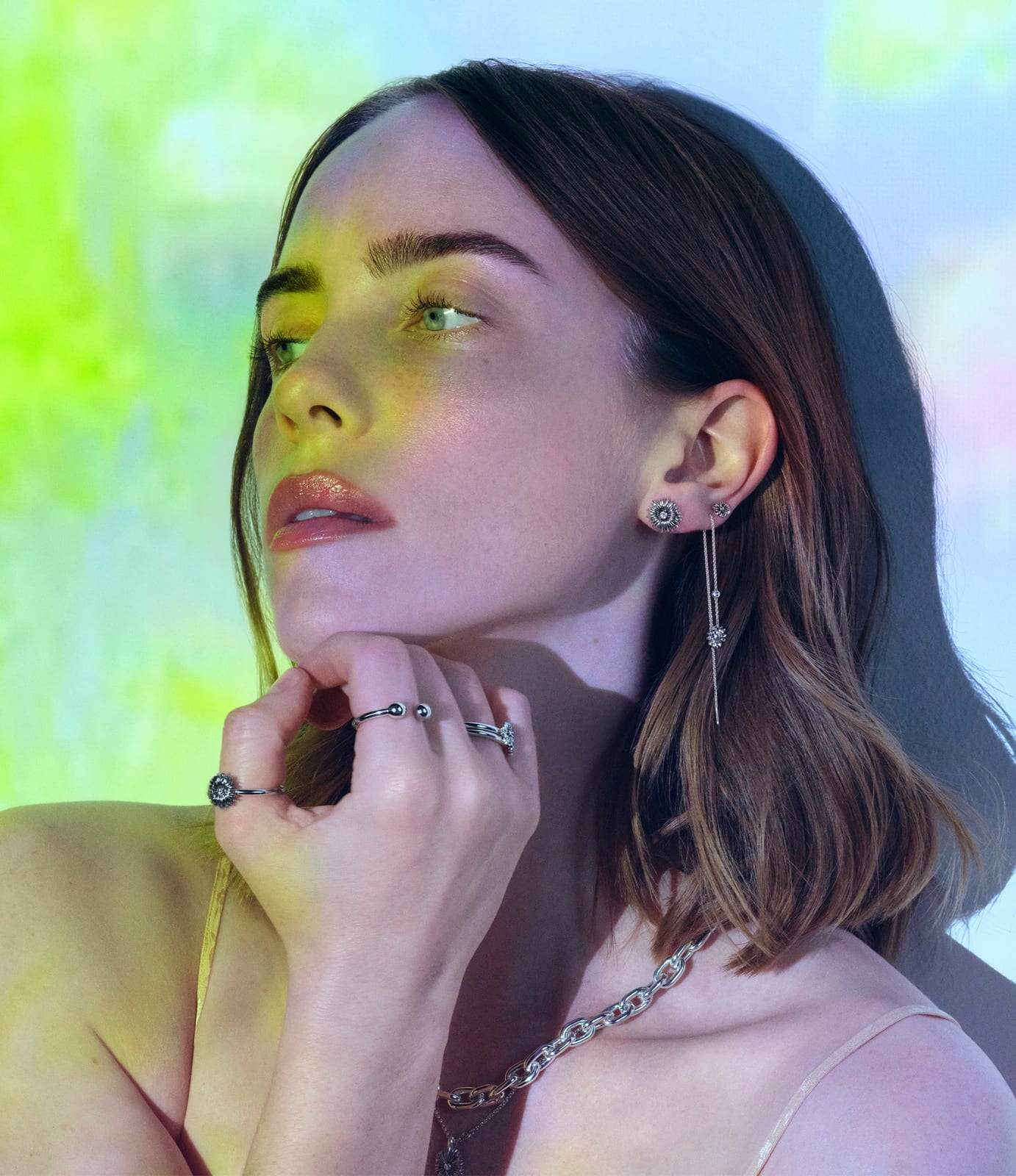 Billie Eilish resting her chin on her hand, wearing a flower stud and a long silver drop-chain earring, silver rings including a flower ring, and a chunky silver link chain over a flower pendant, under green, yellow and lavender projected light