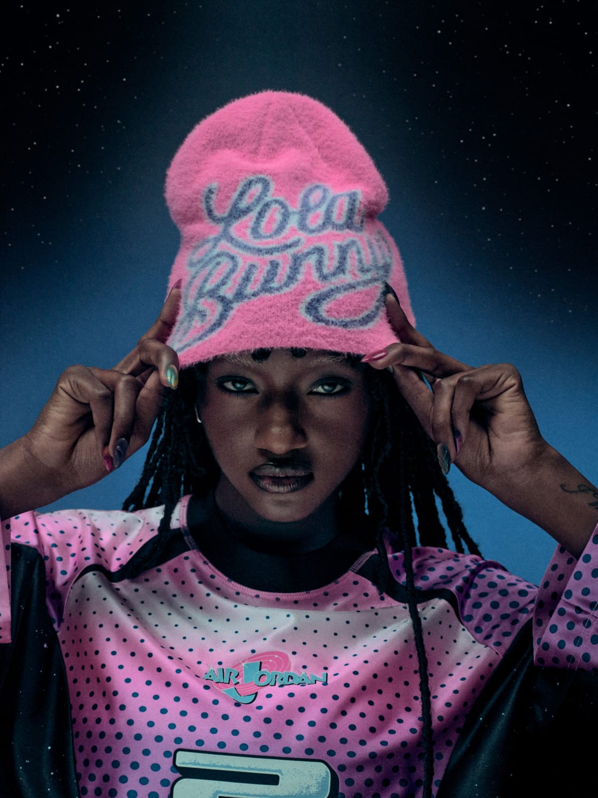 A model in a pink Lola Bunny knit beanie and a pink polka-dot long-sleeve jersey with an Air Jordan Space Jam logo, against a starry blue backdrop
