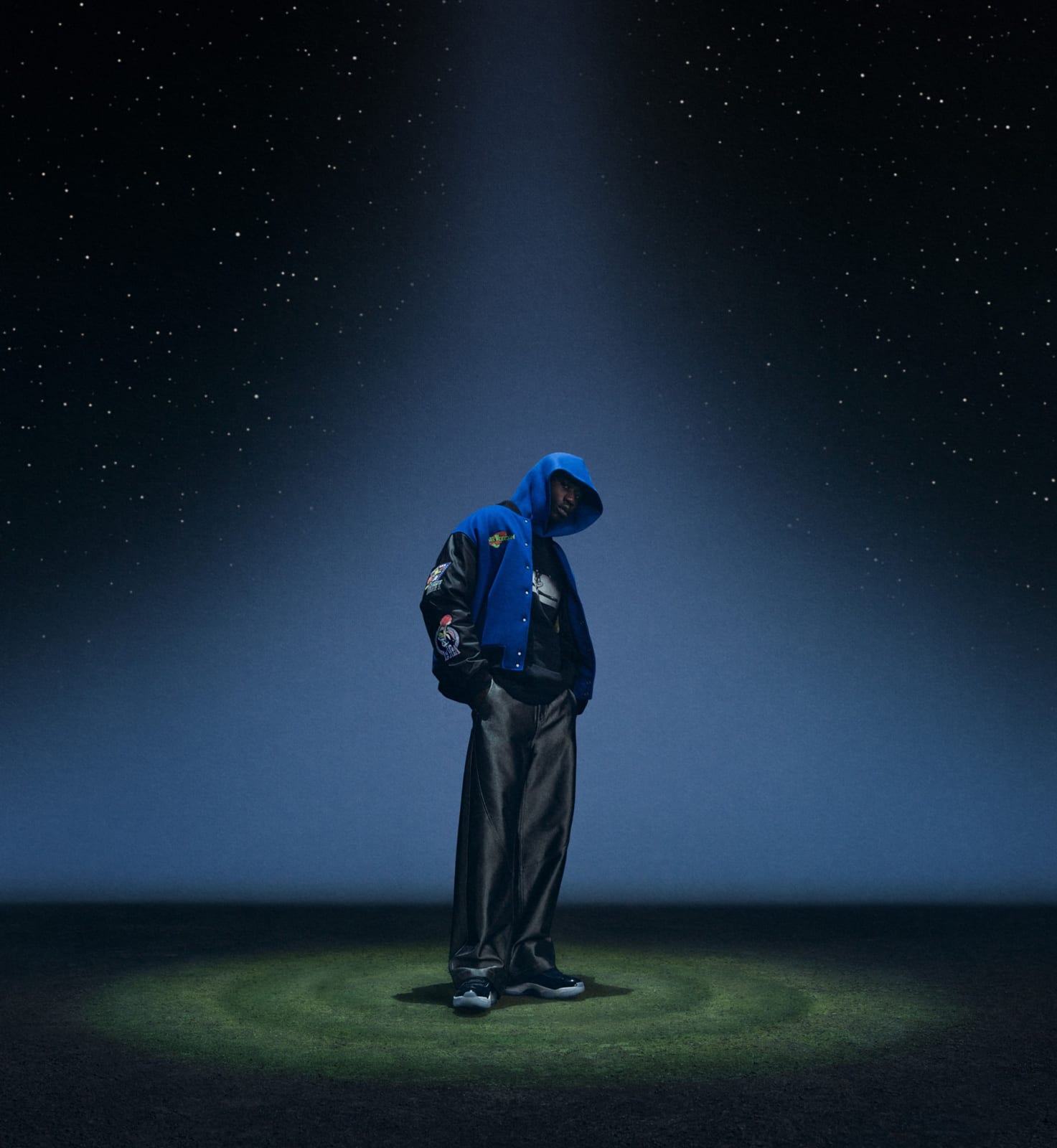 A model in a royal blue faux-wool varsity jacket with black sleeves and Space Jam patches, a blue hood up underneath, standing in a spotlight on a starry night field