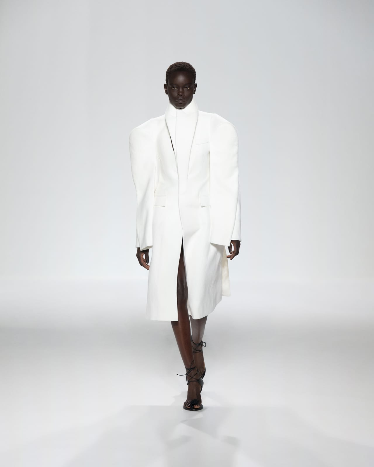 A Jean Paul Gaultier Spring 2027 look: a white knee-length coat with exaggerated rounded shoulders, a raised collar and a cut-out at the chest, worn with black lace-up sandals