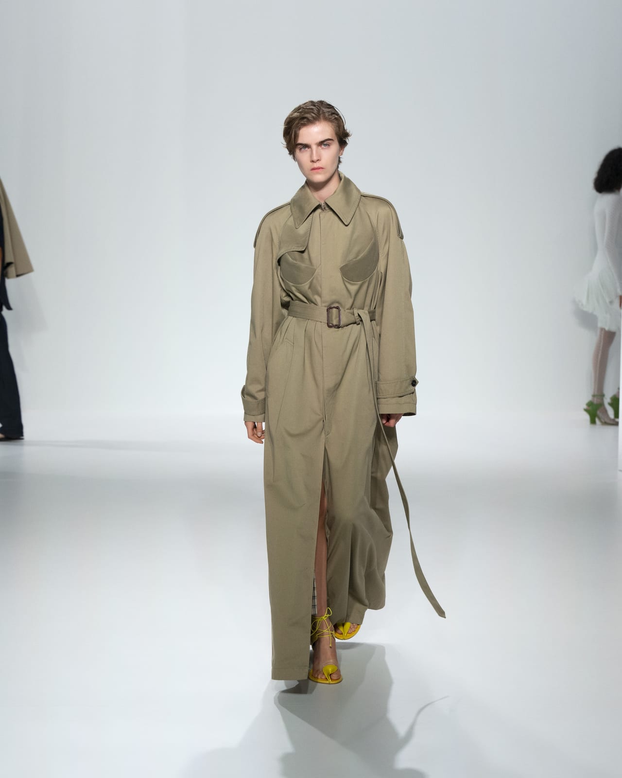 A Jean Paul Gaultier Spring 2027 look: a long khaki belted trench-style coat with rounded cut-out panels at the chest, worn with yellow sandals