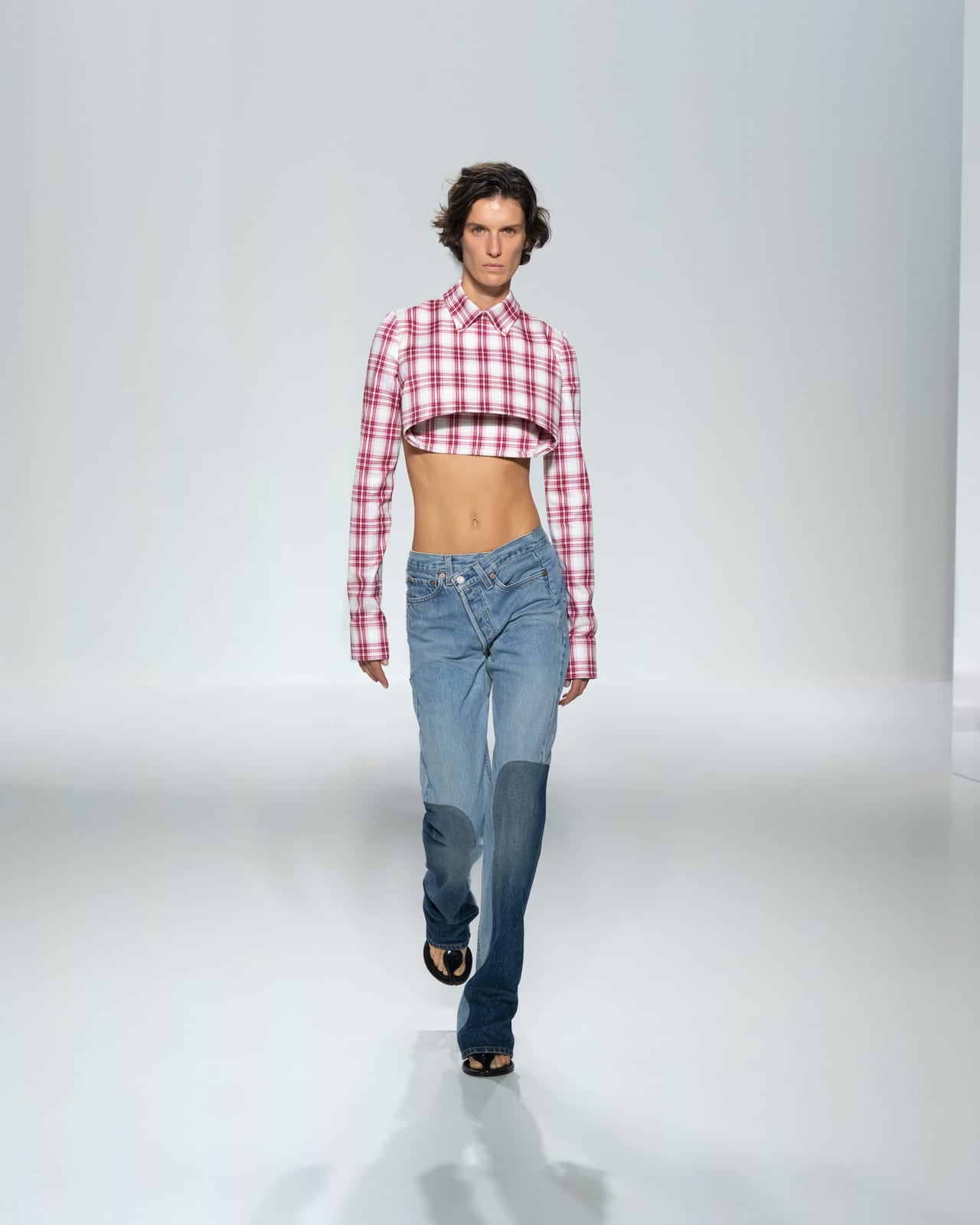 A Jean Paul Gaultier Spring 2027 look: a cropped pink and white check shirt layered over a shorter cropped layer, worn with low-rise jeans with darker knee panels