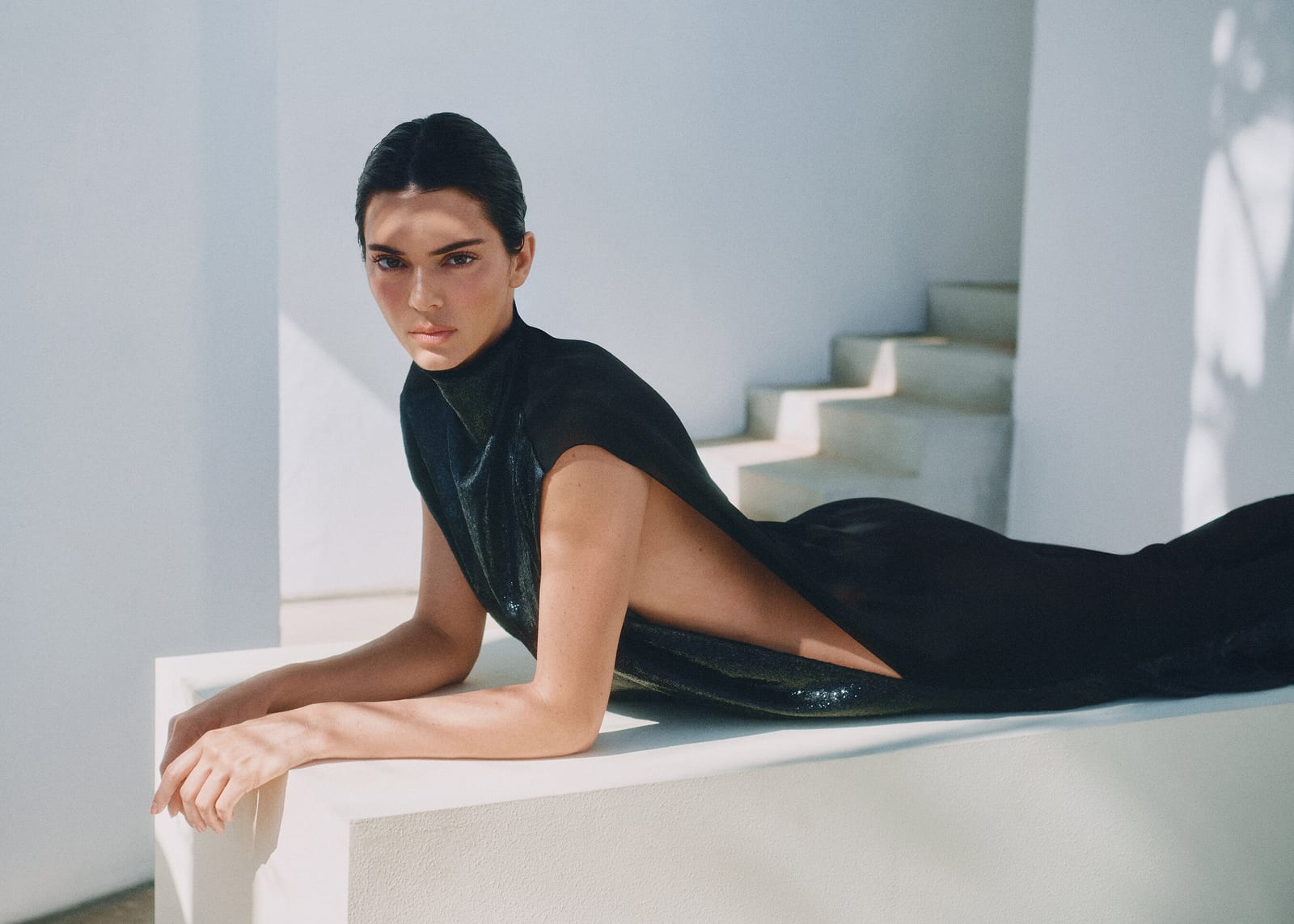 Kendall Jenner, in the photo REVOLVE Group released with the announcement, lying along a white stone ledge in a sleek black high-neck dress with a bare back, white steps and a sunlit wall behind her