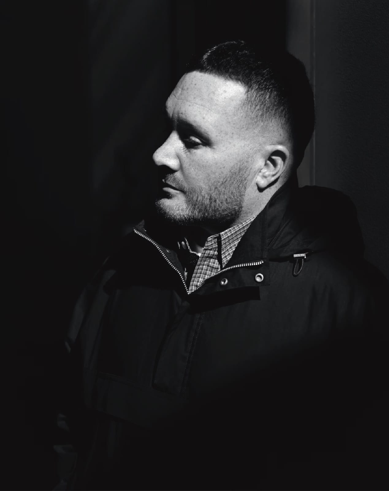 A black-and-white portrait of Kim Jones in profile, half in shadow, wearing a dark hooded parka zipped over a small check shirt