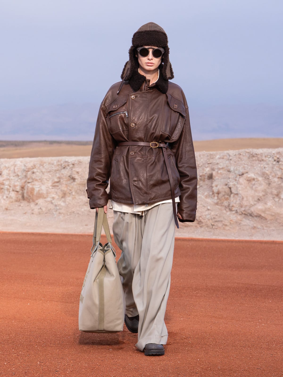 A model in a belted dark-brown leather jacket, a shearling-lined leather trapper hat, sunglasses and wide grey trousers, carrying a large canvas tote on the desert runway