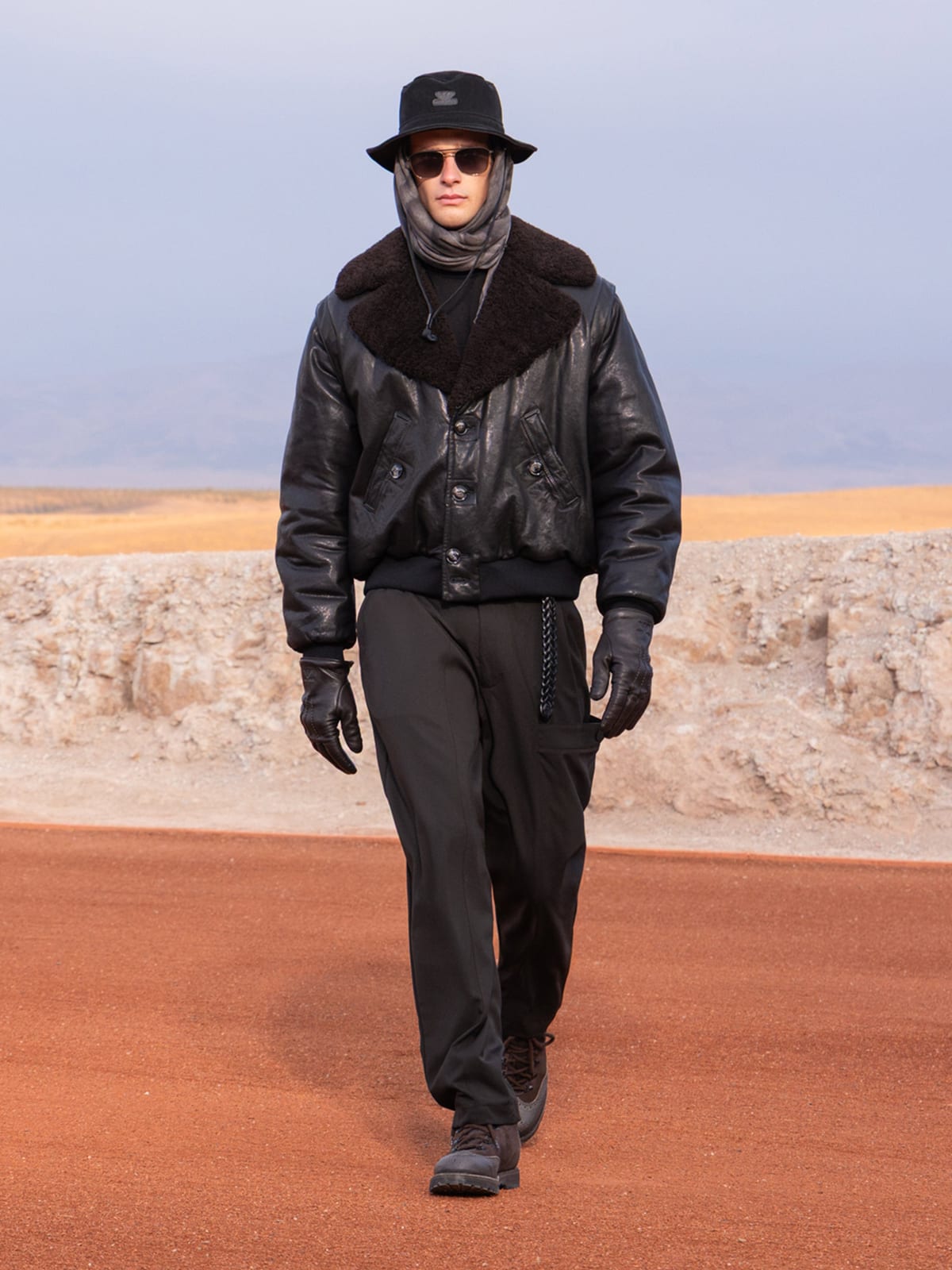 A model in a black leather jacket with a wide black shearling collar, a black bucket hat, sunglasses, a grey scarf over the head, black gloves and black trousers walks a red runway with pale desert rock behind