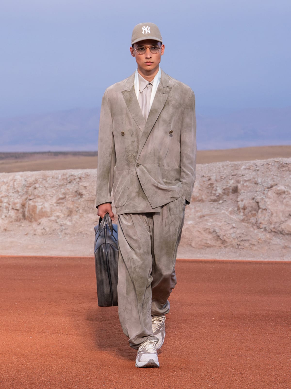 A model in a washed grey-green double-breasted suit with loose trousers, a beige New York Yankees cap, glasses and white sneakers, carrying a dark bag on the desert runway