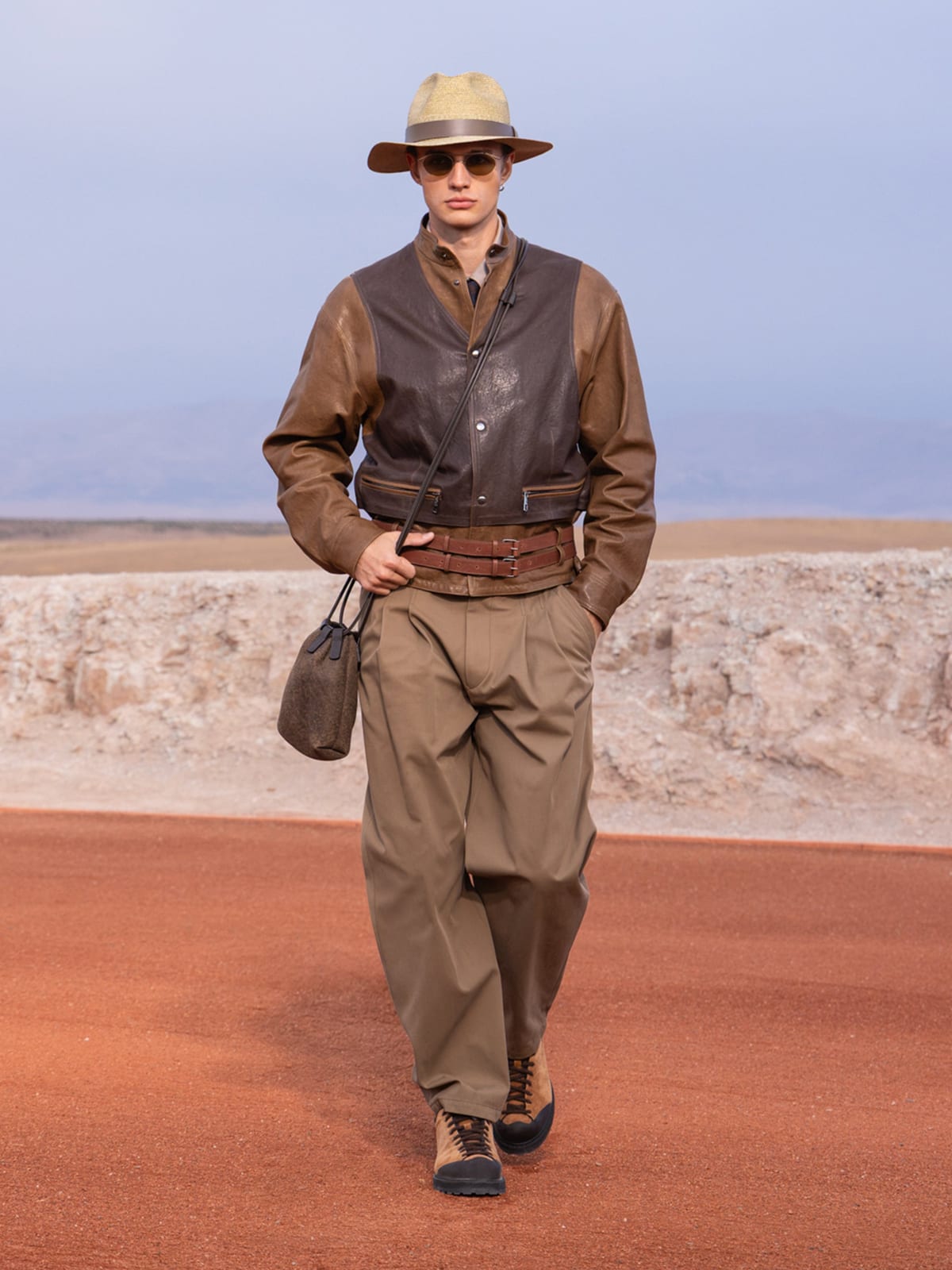 A model in a straw hat and sunglasses walks a red-earth runway in the desert in a two-tone brown leather jacket with darker panels at the chest, a wide double leather belt and pleated khaki trousers, carrying a small leather pouch