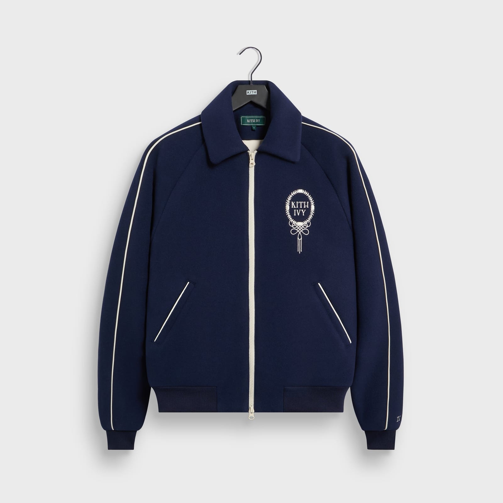 The Kith Ivy wool varsity jacket in Nocturnal navy, with cream piping down the sleeves, a cream two-way zip and a Kith Ivy laurel crest embroidered on the chest, on a Kith hanger