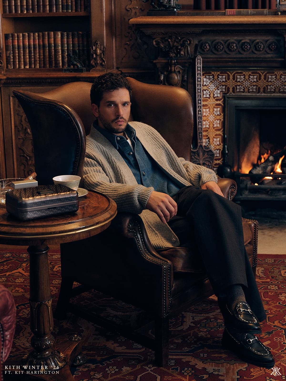 Kit Harington seated in a brown leather wingback chair in a wood-panelled library with a lit fireplace, in a chunky oatmeal cardigan over a denim shirt, dark trousers and black loafers, a cup and saucer and a small dark case on a round table beside him