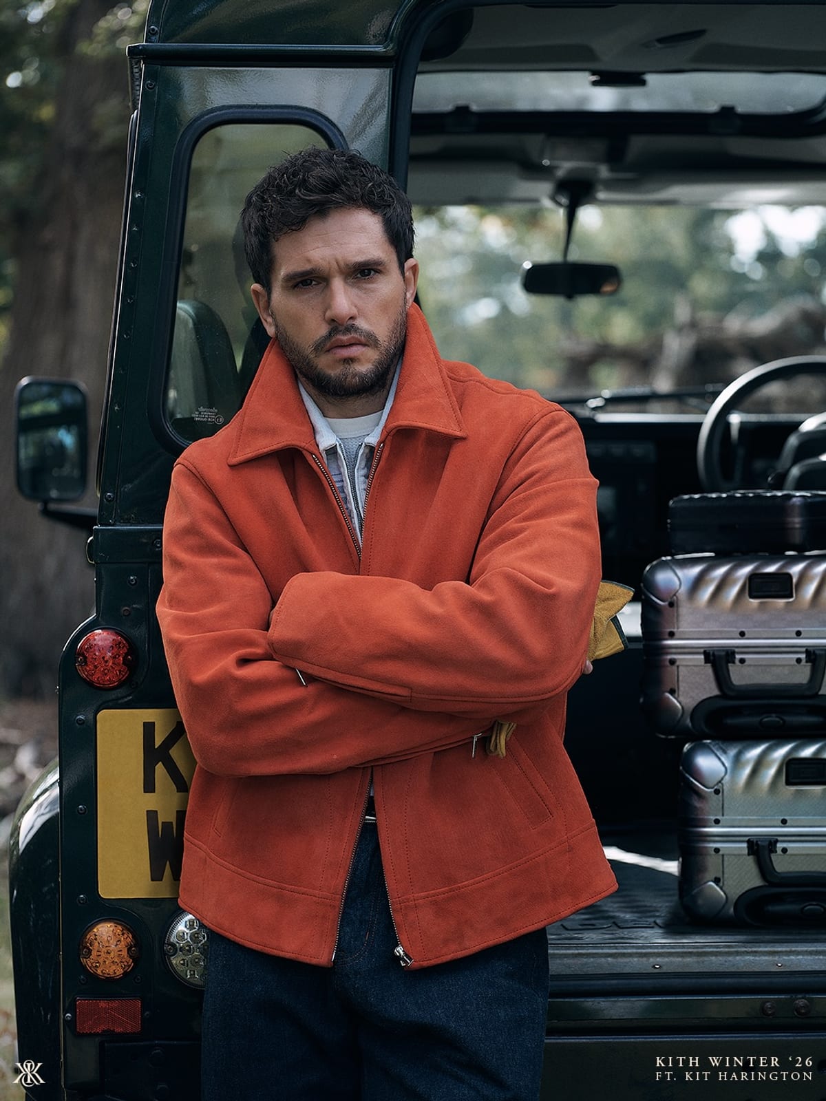 Kit Harington with his arms crossed in a bright orange suede zip jacket over a pale blue shirt, standing in front of the open back of a dark green off-road vehicle with silver aluminium suitcases inside