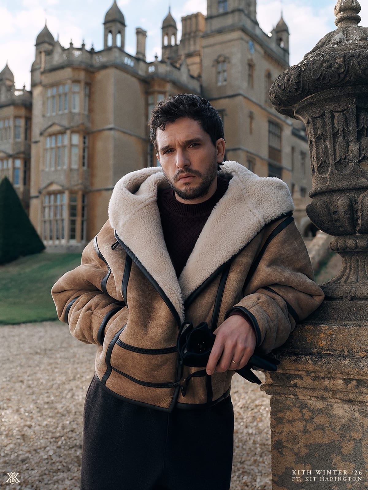 Kit Harington leaning against a carved stone urn in front of a honey-coloured stone manor house with domed towers, wearing a tan shearling-lined hooded jacket with black leather trim over a dark burgundy knit