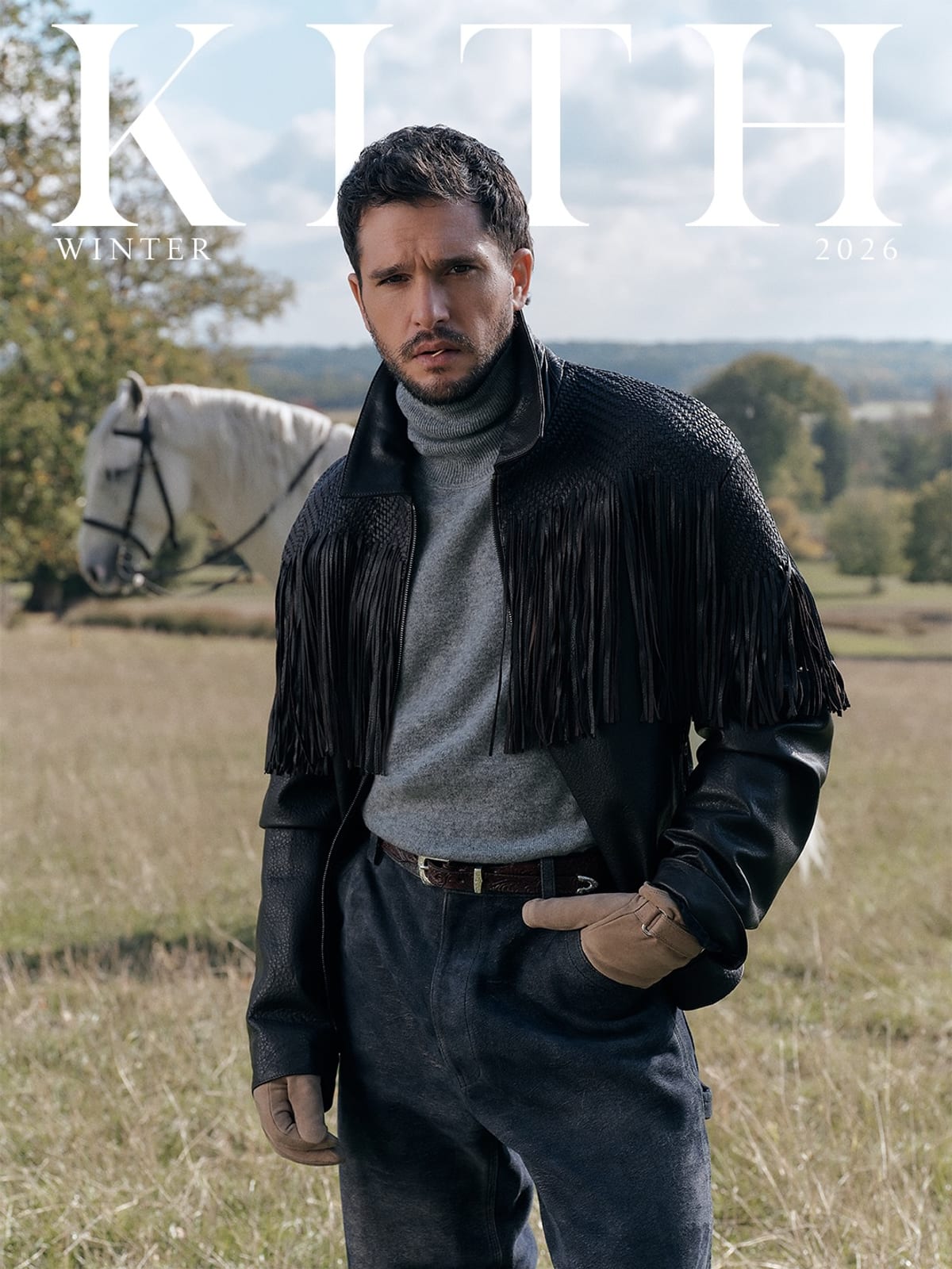 Kit Harington in a black fringed leather jacket over a grey roll-neck, tan gloves on, one hand in his pocket, standing in a field with a white horse behind him, under the large white KITH Winter 2026 wordmark