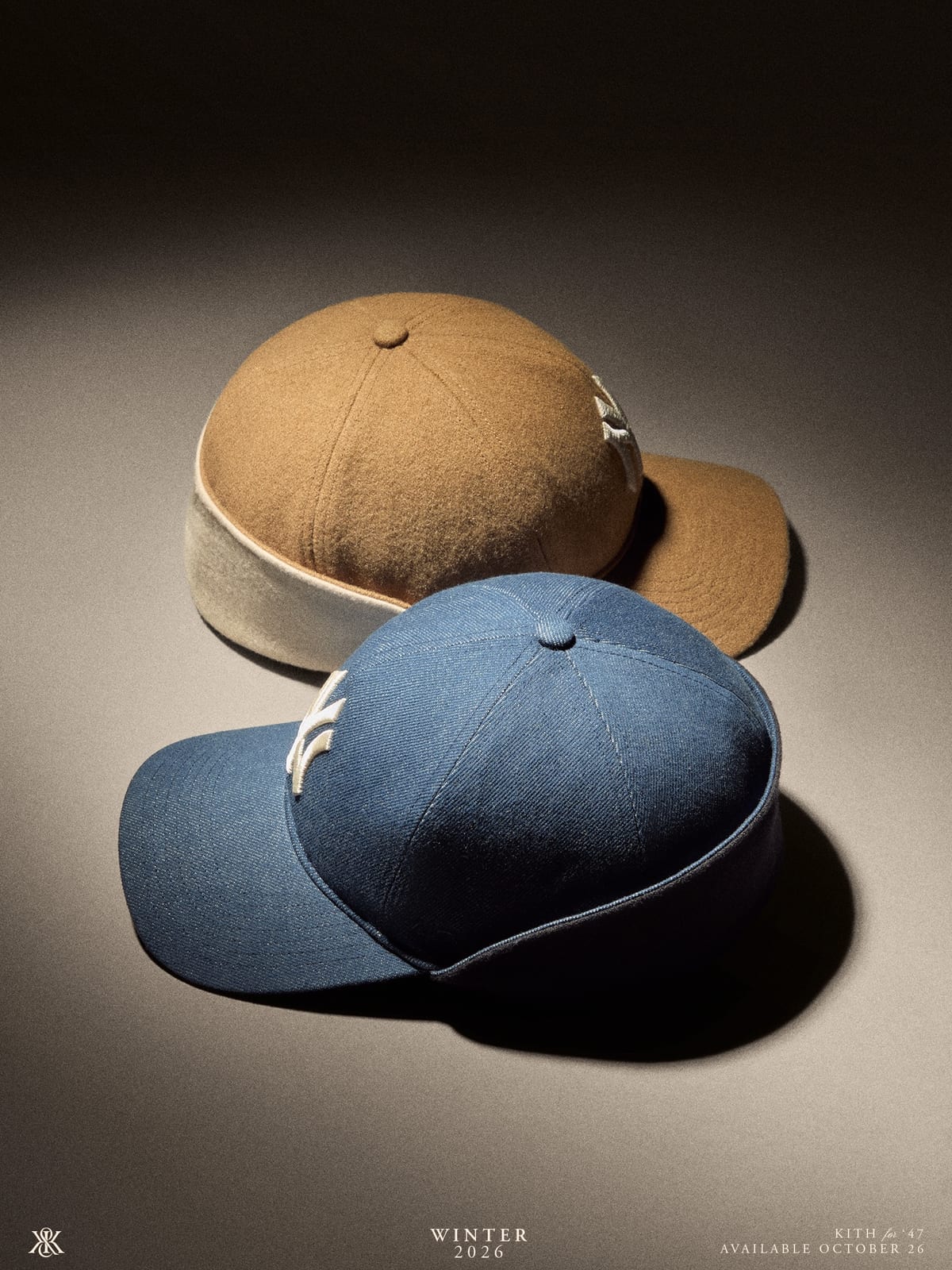Two Kith for '47 baseball caps with the white New York Yankees logo, a tan wool one with a cream band behind a blue one, against a grey backdrop