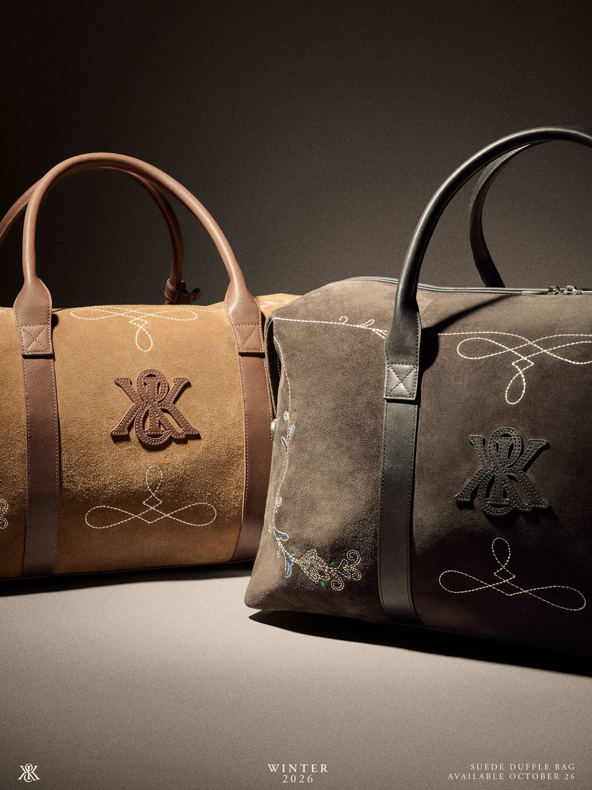 Two suede duffle bags side by side, tan on the left and dark grey-brown on the right, each with leather handles, a leather Kith crest patch and Western scrollwork stitched on the front, against a grey backdrop
