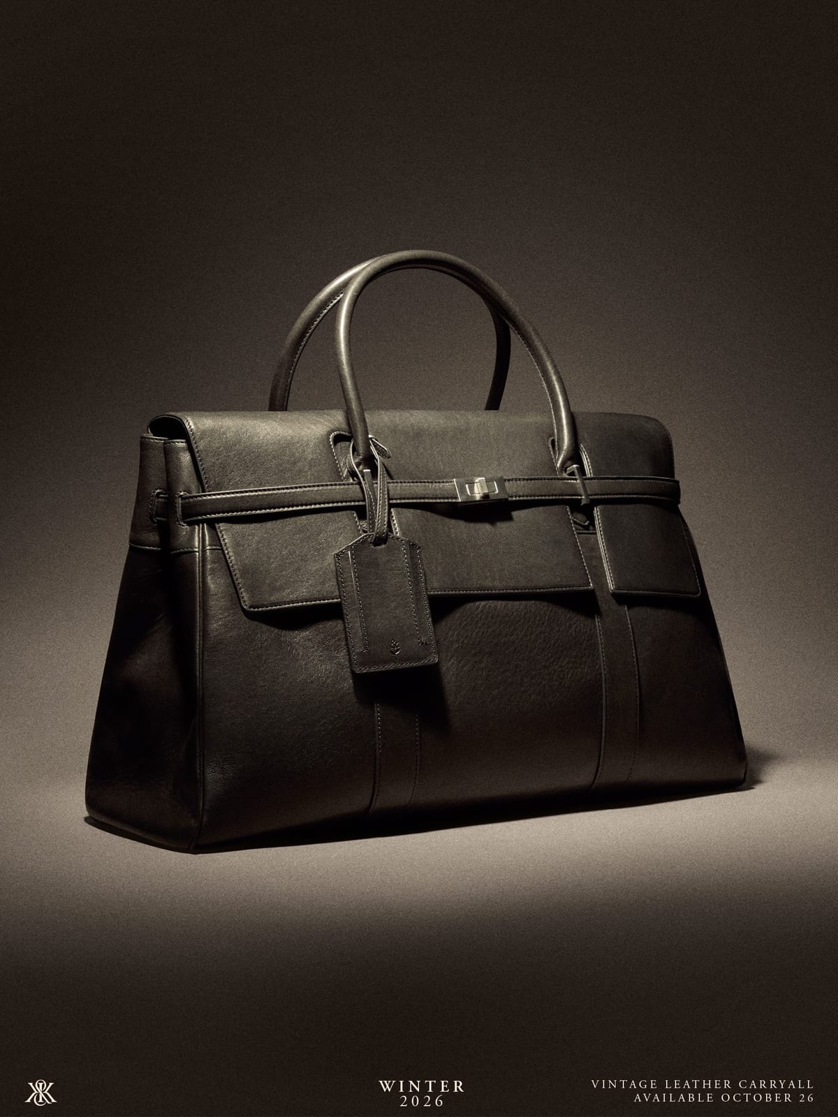A black leather carryall bag with two top handles, a front flap with a strap closure and a hanging leather luggage tag, lit from the side against a dark grey backdrop, labelled Vintage Leather Carryall, Available October 26