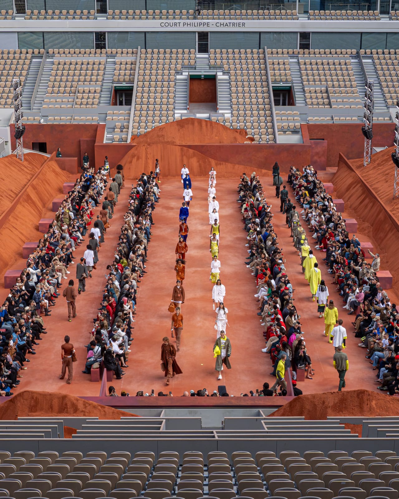 Lacoste's Spring 2027 show seen from the stands of Court Philippe-Chatrier at Roland-Garros: models walking a clay runway between rows of guests, with banks of clay piled around the court