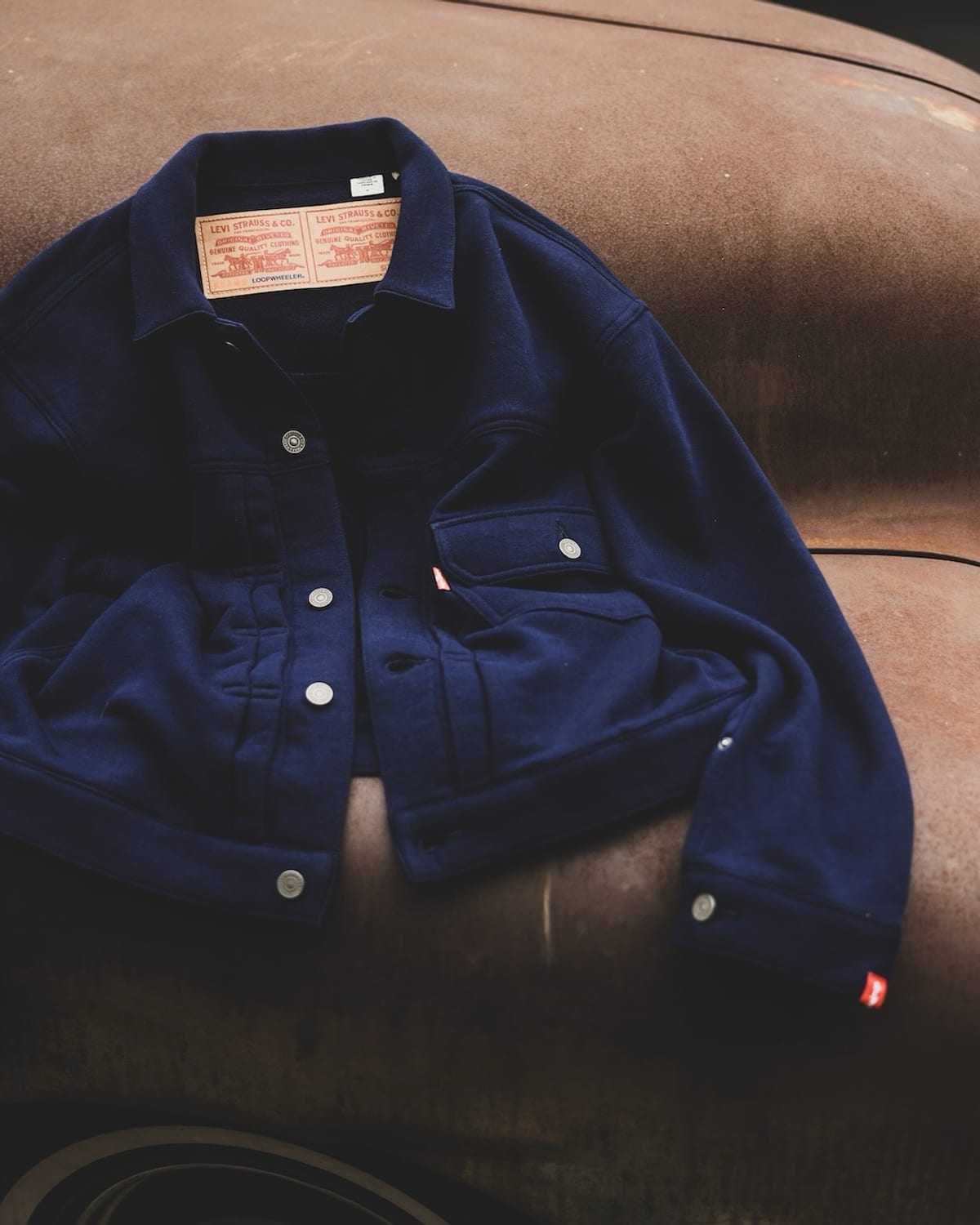 A navy Levi's x BEAMS x LOOPWHEELER Super Wide Trucker in sweatshirt fabric, laid flat on the rusted body of an old car, with a leather-look patch inside the collar, silver buttons and small red tabs