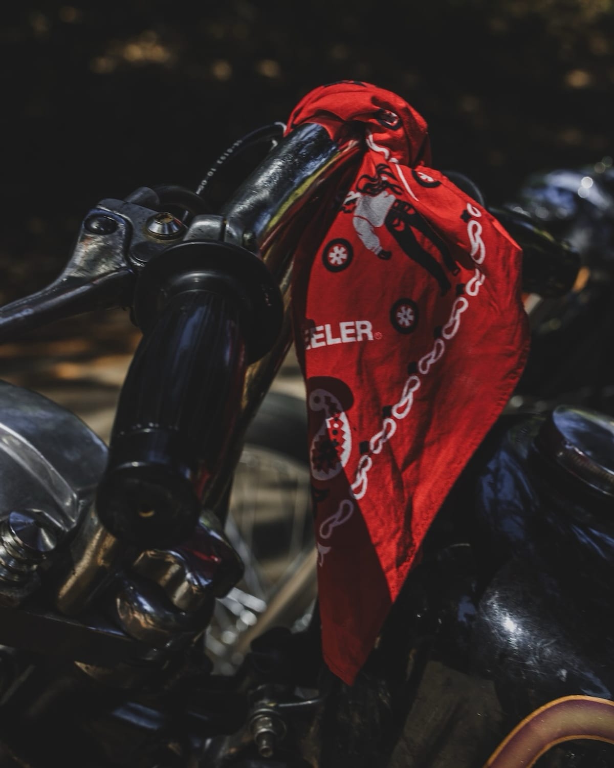 A red bandana with a white chain and figure print, and LOOPWHEELER lettering, tied around a motorcycle's handlebar