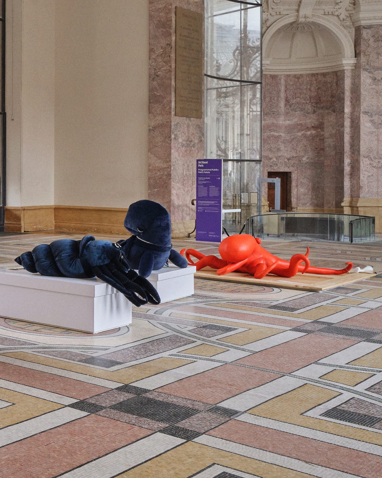 A dark blue velvet hermit crab sculpture on a white plinth and a red leather octopus sprawled on a plywood board, on the Petit Palais's mosaic floor, with a purple Art Basel Paris public program sign behind them