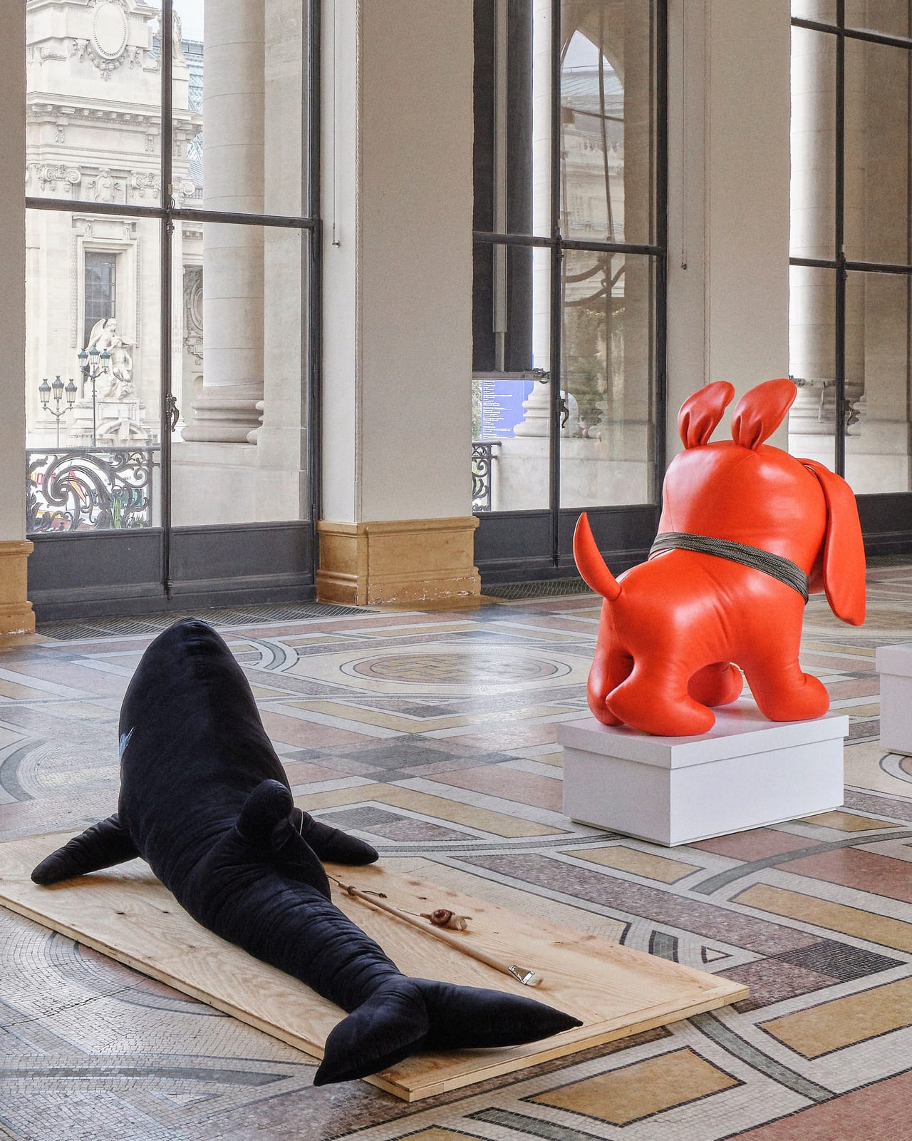 A large black plush whale lying on a plywood board on the Petit Palais's mosaic floor, beside an orange leather dog sculpture on a white plinth, in front of tall windows