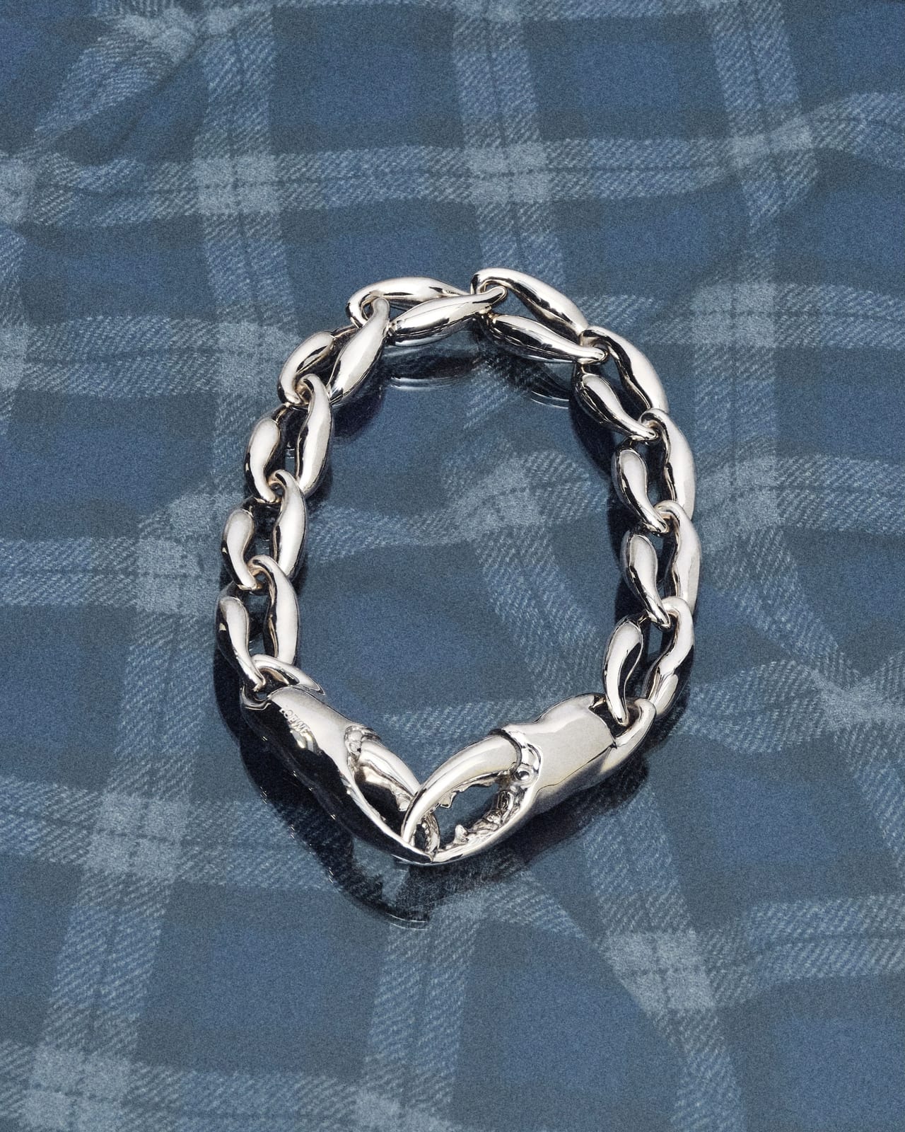 A chunky silver chain bracelet whose clasp is shaped like a pair of pincer claws, lying on blue and grey tartan fabric