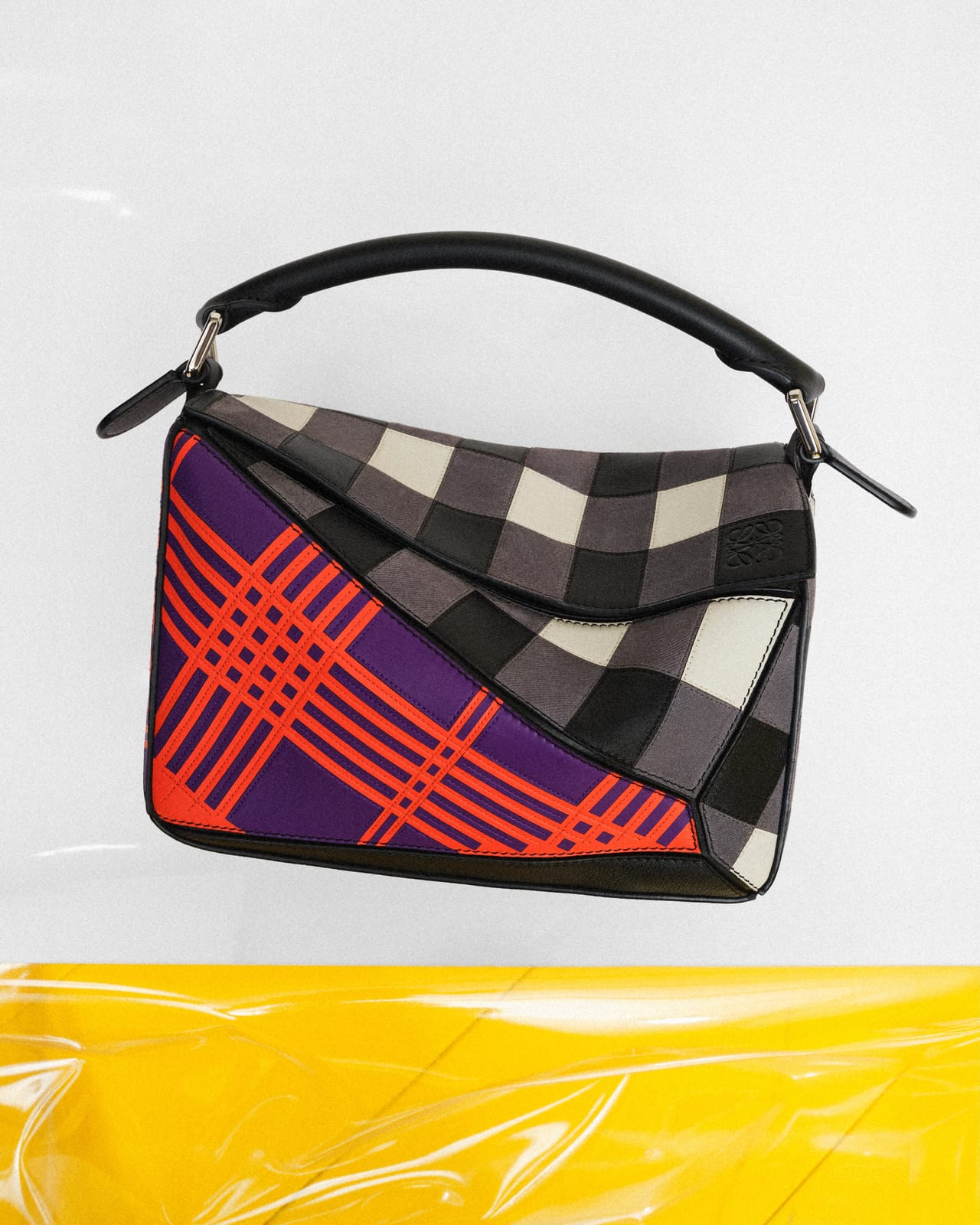 A Loewe handbag with a black leather top handle, made of angular panels in grey-and-white check and orange tartan on purple, against a white backdrop above yellow plastic sheeting