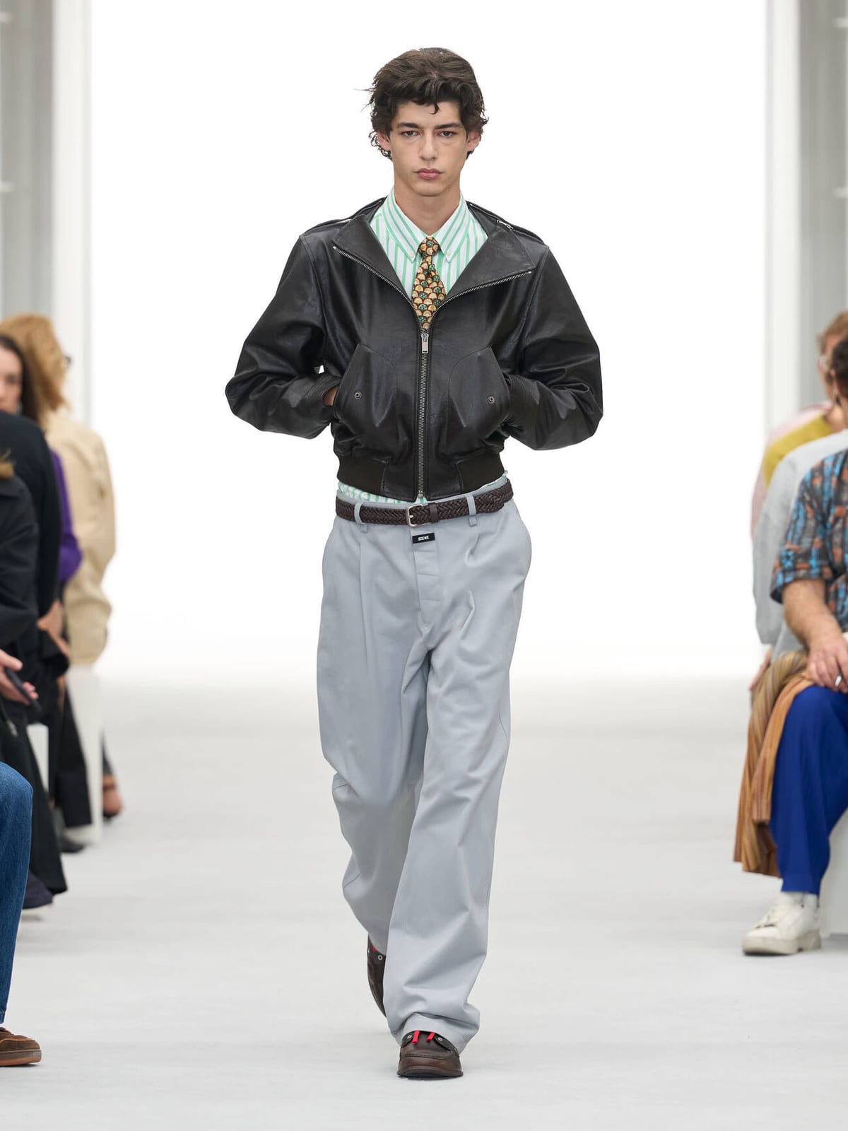 A model on Loewe's white runway in a cropped black leather bomber jacket, a striped shirt with a patterned tie, grey trousers and a belt