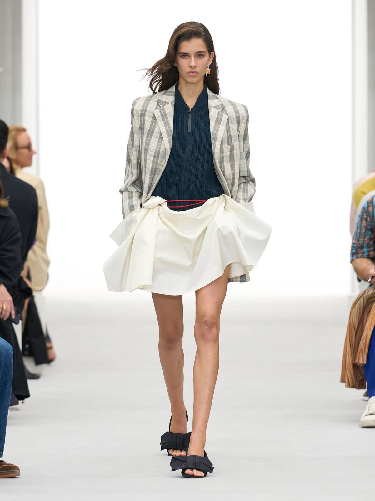 A model in a grey check blazer over a navy zip top and a short white skirt with a flared, gathered hem, with black bow sandals