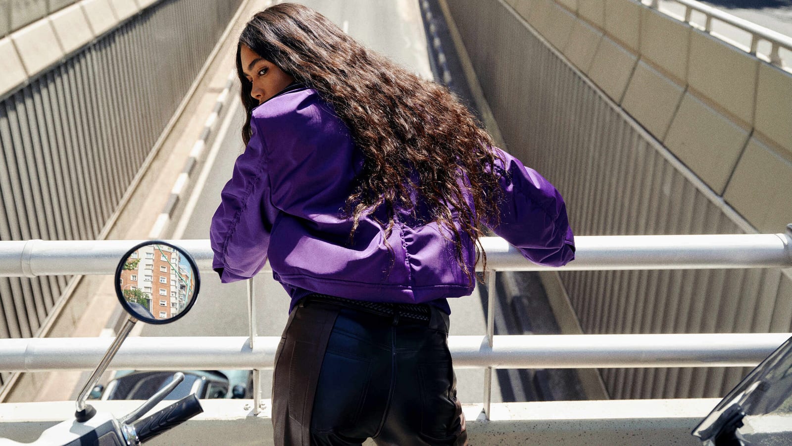 A woman with long curly dark hair, seen from behind and looking back over her shoulder, in a cropped purple bomber jacket and black leather trousers, leaning on a metal railing above a sunken road between concrete walls, a scooter's round mirror reflecting an apartment block beside her