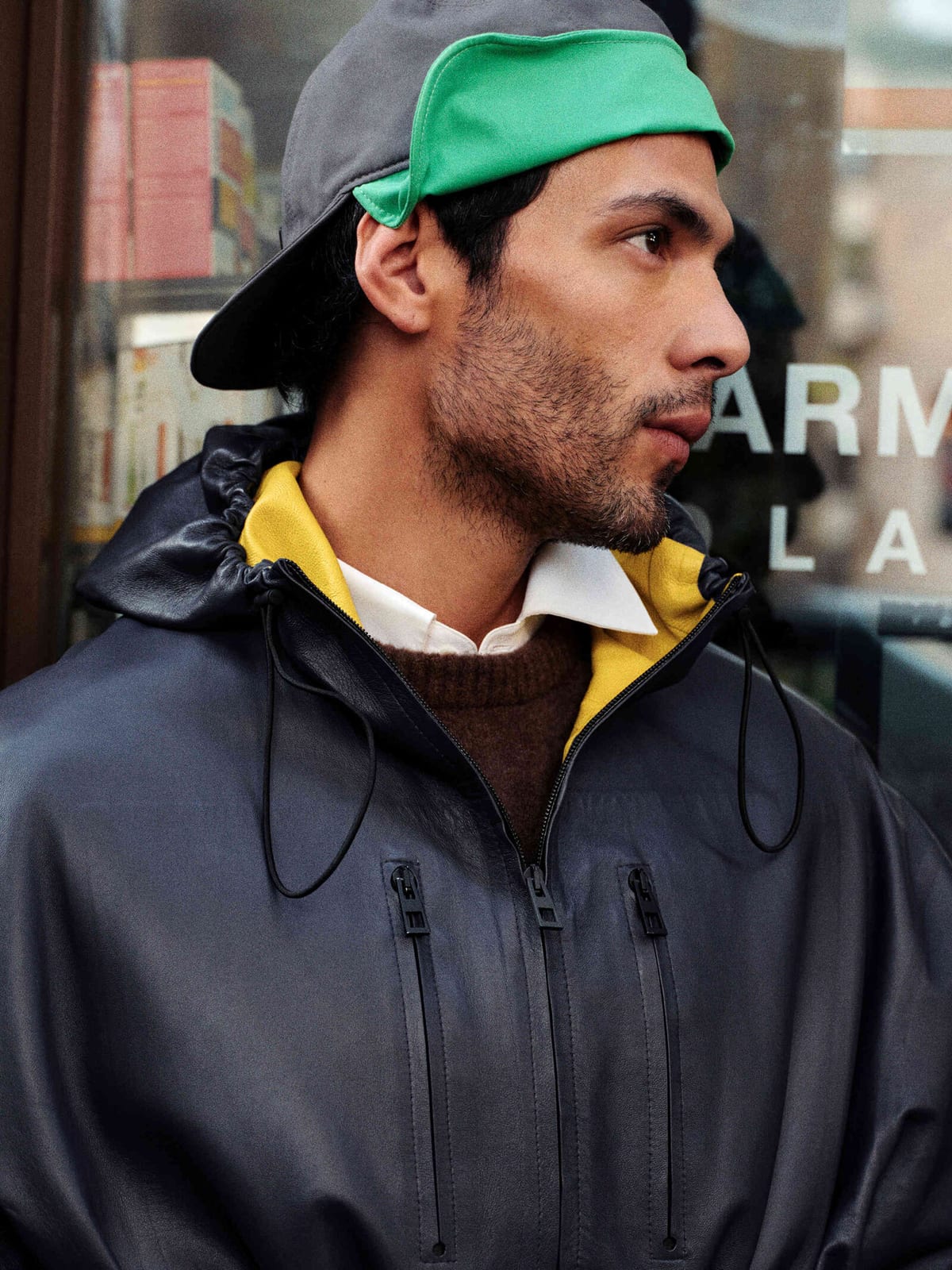 A man in profile in front of a shop window, wearing a grey cap turned backwards with a green back panel showing, a navy leather jacket with a drawstring hood lined in yellow leather, zip pockets on the chest, over a brown knit and a white shirt collar