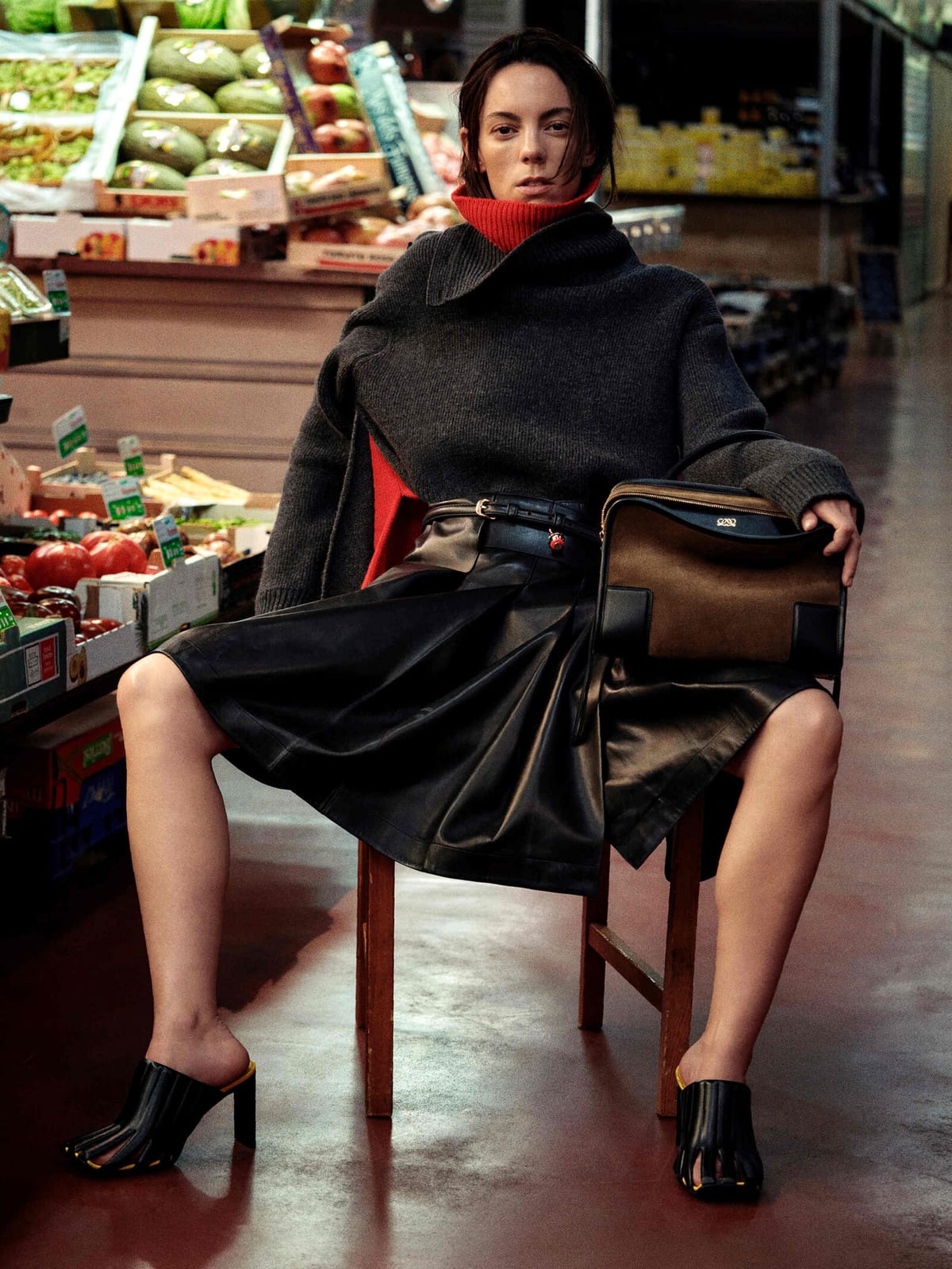Victoria Luengo seated on a wooden chair in the aisle of a fruit and vegetable market, in a charcoal knit with an asymmetric wrapped collar over a red roll-neck, a full black leather skirt with a belt, black caged heeled mules, and a brown suede and black leather top-handle bag with a gold Anagram on her lap