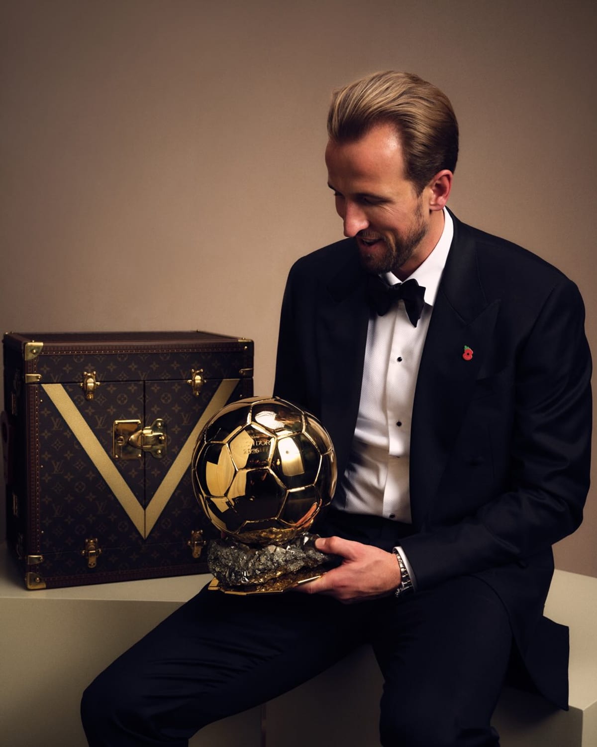 Harry Kane in a black tuxedo and bow tie with a red poppy pin, smiling down at the golden Ballon d'Or trophy he holds, beside a brown Louis Vuitton Monogram trunk with a gold V on its front